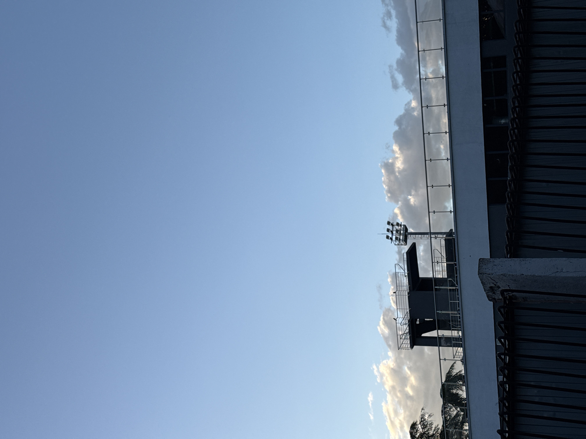 The image shows a building with a tall structure on top, likely a sports arena or stadium lighting rig. The structure is made of metal and has multiple lights mounted on it. The sky is visible in the background, with some clouds present. The building has a white exterior with glass railings. The perspective is from below, looking up at the building.