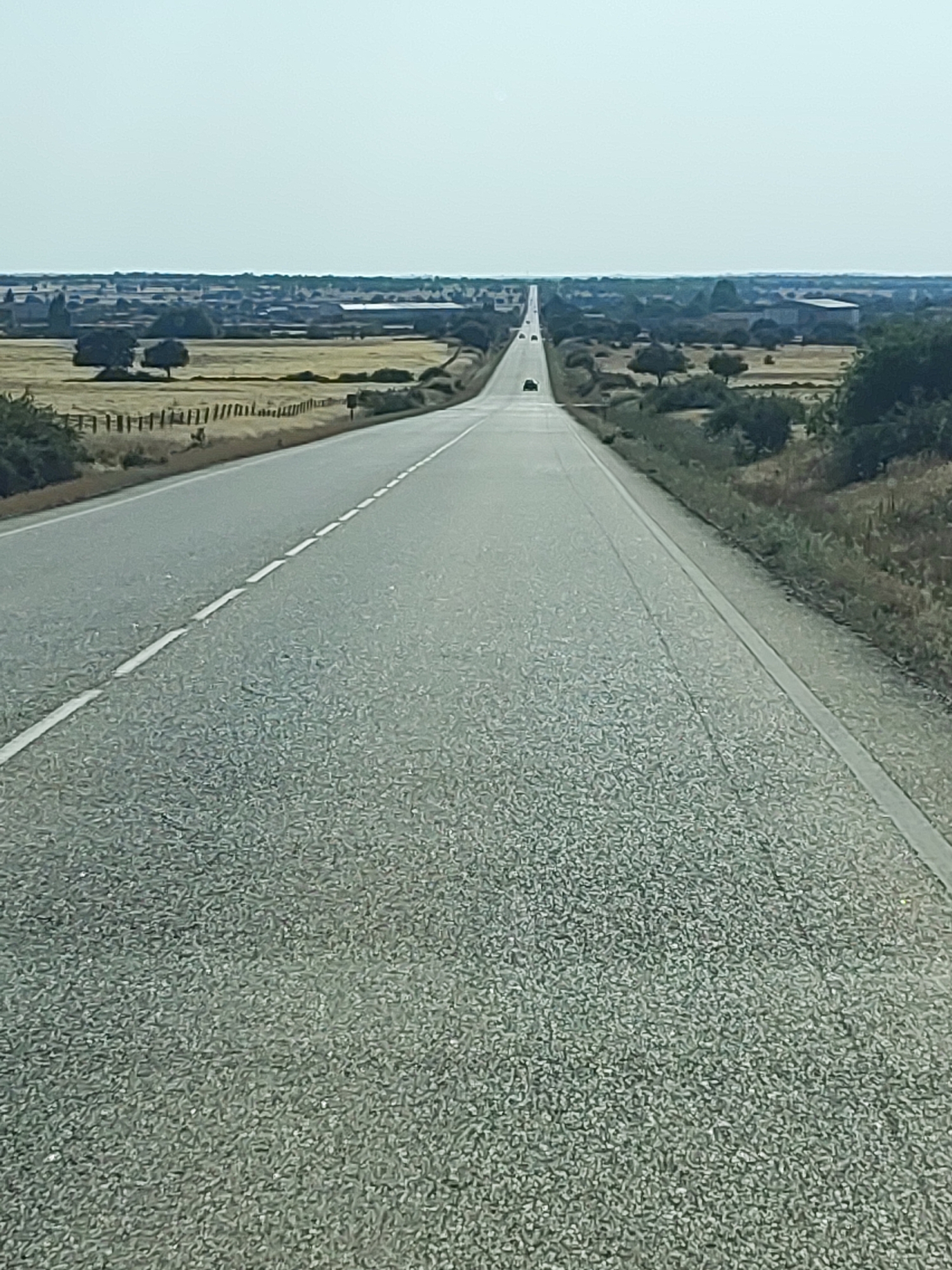 The image shows a long, straight road disappearing into the horizon in a rural landscape. The road is paved with asphalt and has white dashed lines marking the lanes. The landscape on either side of the road consists of dry fields, scattered trees, and some distant buildings, possibly farms or a small village. The sky is a pale, washed-out blue, indicating a clear, possibly hot, day. A few dark-colored vehicles are visible on the road in the distance, suggesting a very sparse traffic. The perspective is from a low angle, as if taken from a vehicle moving forward on the road. The scene evokes a sense of openness and vastness typical of rural Spain. The location is identified as being in Villar de Peralonso, Spain, which is a municipality in the province of Salamanca, in the autonomous community of Castile and León.