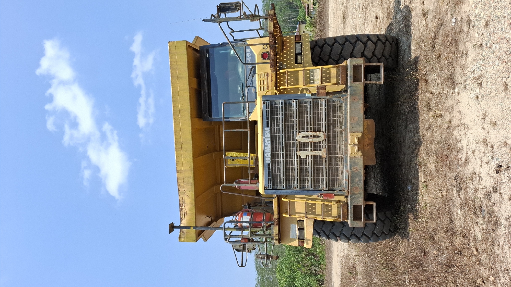 An extreme wide shot captures a large, yellow Komatsu dump truck from a frontal, slightly low-angle perspective in a dry, outdoor environment. The truck dominates the frame, its massive tires and imposing grill with the number "10" prominently displayed. The cab, with its tinted windows, is visible at the top right of the grill. Various safety features, such as railings, ladders, and fire extinguishers, are attached to the truck's exterior. The background consists of sparse, dry vegetation, sandy terrain, and a clear blue sky with scattered white clouds. The overall scene suggests an industrial or mining setting, likely in a location with arid conditions. The specific city of Unknown, Indonesia, is an assumption about the location and not directly verifiable from the image content.