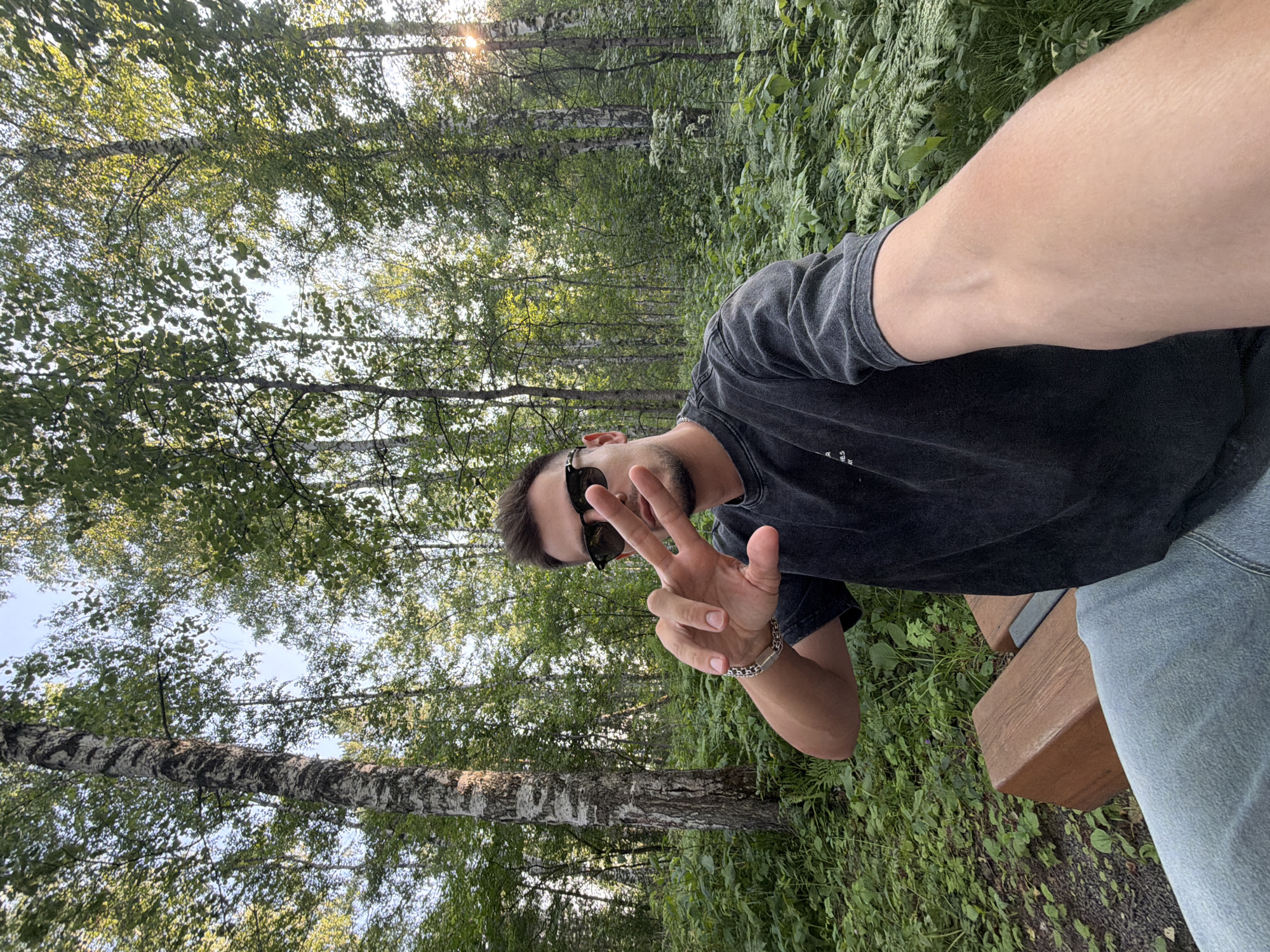 A man is taking a selfie in a forest. He is wearing a dark grey t-shirt, light blue jeans, and black sunglasses. He is sitting on a wooden bench and making a peace sign with his right hand. The background consists of tall birch trees and lush green foliage. The lighting suggests it is late afternoon. The specific location within Krasnoyarsk, Russia, cannot be determined from the image alone.