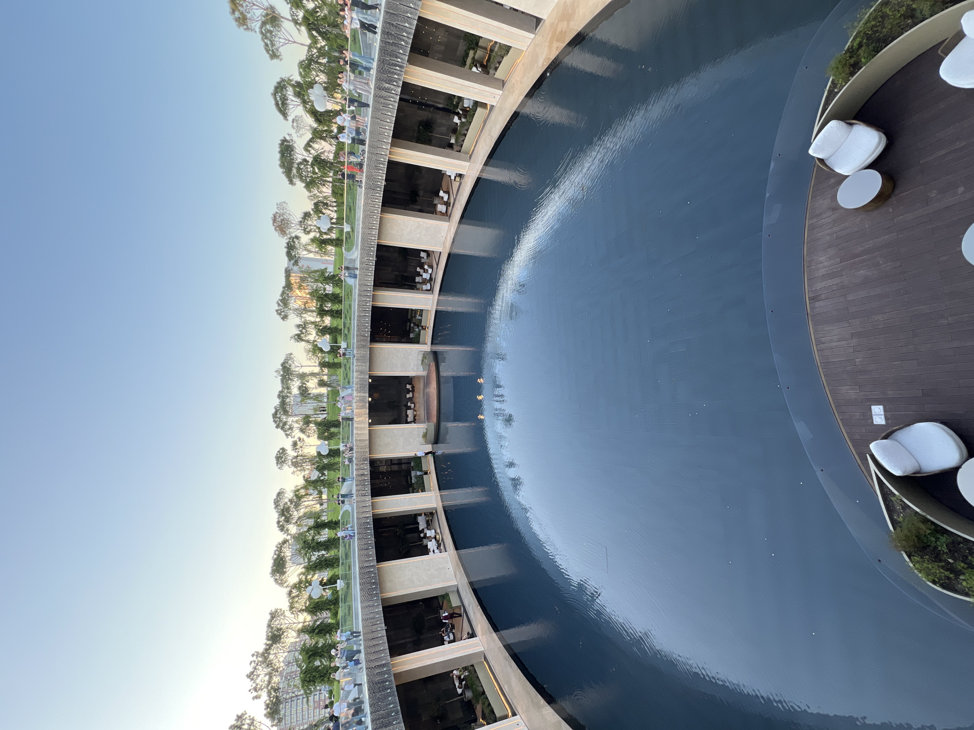 This image captures a modern architectural and landscape design in Krasnodar, Russia, likely during the late afternoon or early evening (golden hour). The view is from an elevated perspective, looking down and across a circular or semi-circular outdoor complex.
In the foreground, a dark wooden deck with a slightly curved edge is visible. Two white cushioned outdoor chairs and a round, light-colored table are placed on the deck, suggesting a lounge or seating area. This deck borders a large, dark blue body of water, possibly a decorative pool or fountain, which occupies a significant portion of the image. The water surface shows gentle ripples and reflects the clear, light blue sky above.
Across the water, a multi-level concrete or stone structure curves along the edge. This building features open-air terraces or balconies on several floors, each supported by pillars and offering views of the water. On these terraces, smaller tables and chairs are arranged, and several people can be seen dining or socializing. The interior lights of these sections are illuminated, adding a warm glow.
Above this structure, a wide promenade or walkway extends into the distance, lined with numerous slender trees resembling pines and modern, spherical streetlights. Many people are seen strolling along this walkway, enjoying the outdoor environment. The background shows more greenery and the faint outlines of distant buildings under a clear, light blue sky that fades towards the horizon.
The overall scene depicts a vibrant public space designed for leisure and social gathering, characterized by contemporary architecture, landscaping, and a prominent water feature. No visible text is present in the image.