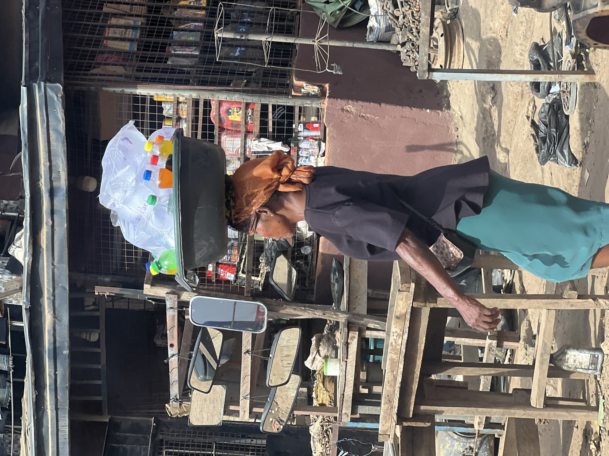 This image depicts an individual engaged in street vending within a bustling outdoor market or street scene in Nkpor, Nigeria, under bright daylight conditions. The person, appearing to be an older woman, is shown in profile, facing to the right. She is carrying a large, black basin on her head, which is filled with several plastic bottles containing various colored liquids (orange, yellow, and green) and clear plastic bags, presumably items for sale. One plastic bag clearly shows the text "ssiPack". She is dressed in a dark long-sleeved top, a teal-green skirt, and a reddish-brown headscarf tied around her head. A small, leopard-print patterned bag hangs from her left arm.
The surrounding environment suggests a busy commercial area. To her right, a series of crude wooden frames and shelves display various goods, prominently featuring multiple vehicle rear-view mirrors. The ground beneath is uneven and dusty. In the background, a dark metal mesh structure acts as a barrier or part of a stall, with shelves behind it stocked with what appear to be various packaged items. Further to the right in the background, discarded metal parts, possibly from vehicles, and a partial view of a bicycle are visible. The strong shadows cast by the structures indicate a bright, sunny day, likely midday or early afternoon. The scene captures a typical moment of commerce and daily life in a vibrant local market.