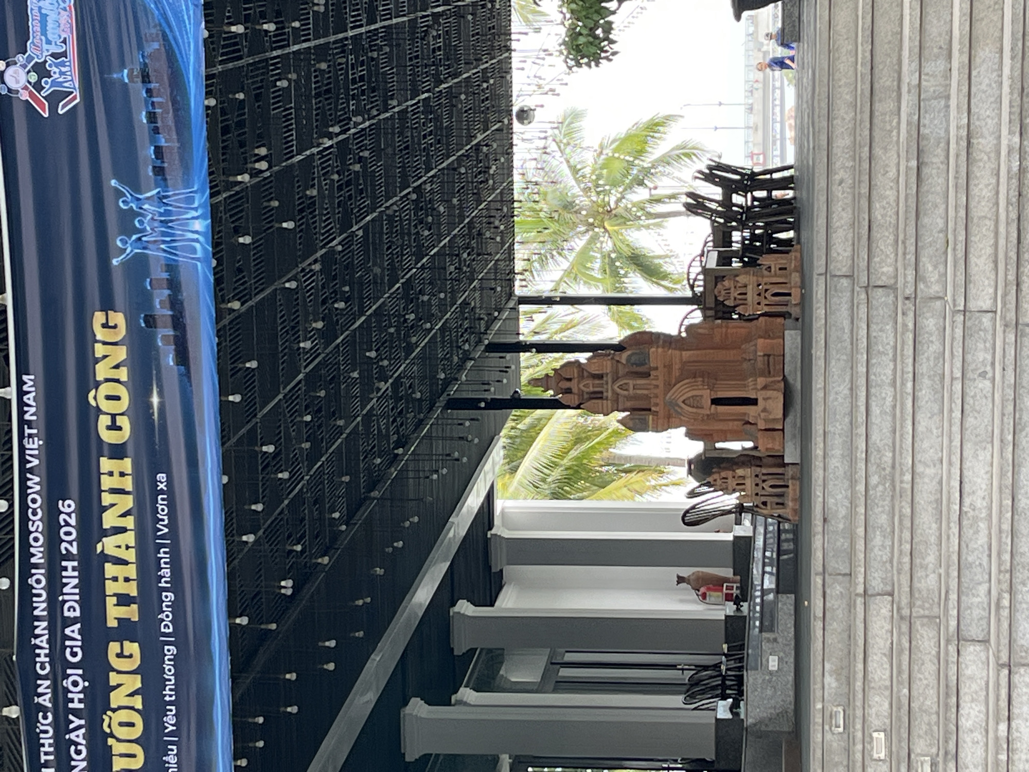 The image shows a covered outdoor seating area, likely a restaurant or event space, with a view of a body of water and a bridge in the distance. The architecture features white columns and a dark, grid-like ceiling with numerous small lightbulbs hanging down. In the foreground, there are wide concrete steps leading up to the seating area.
On a raised platform within the seating area, there are several dark chairs arranged around tables, and two ornamental terracotta structures resembling ancient buildings or pagodas. A tall, narrow vase and a red fire extinguisher are also visible on the platform.
In the middle ground, palm trees are visible through the open sides of the structure, suggesting a tropical or subtropical climate. The sky is bright, indicating daylight.
In the background, people are visible near the water's edge. One person in blue is standing, and another smaller figure is also present. A bridge spans the water in the far distance.
The banner at the top of the image, which is mostly obscured by the ceiling, has text in Vietnamese. Partially visible phrases include "THỨC ĂN CHĂN NUÔI MOSCOW VIỆT NAM" (Moscow Vietnam Livestock Food), "NGÀY HỘI GIA ĐÌNH 2026" (Family Festival 2026), and "VƯƠNG THÀNH CÔNG" (Conquering Success). There is also a logo with stylized figures and the word "Family". The presence of this banner strongly suggests the location is within Nha Trang, Vietnam, given the Vietnamese language used. The context of a "Family Festival 2026" and "Moscow Vietnam Livestock Food" implies this could be a promotional event or an establishment related to food and family gatherings.