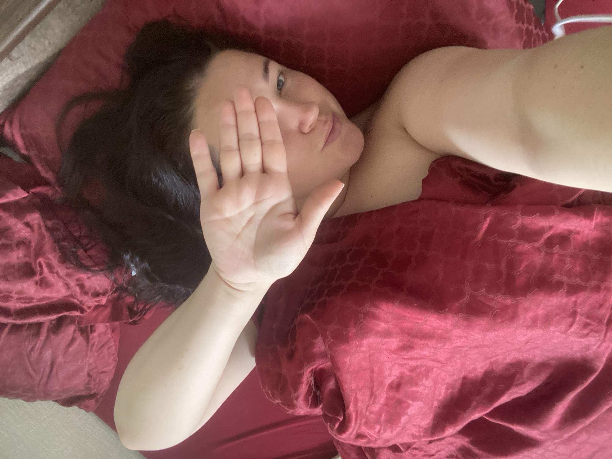 A woman lies in a bed covered with red sheets. She holds her hand up, obscuring part of her face. Her dark hair is spread out on a red pillow. The location is not discernible as being in Sochi, Russia.