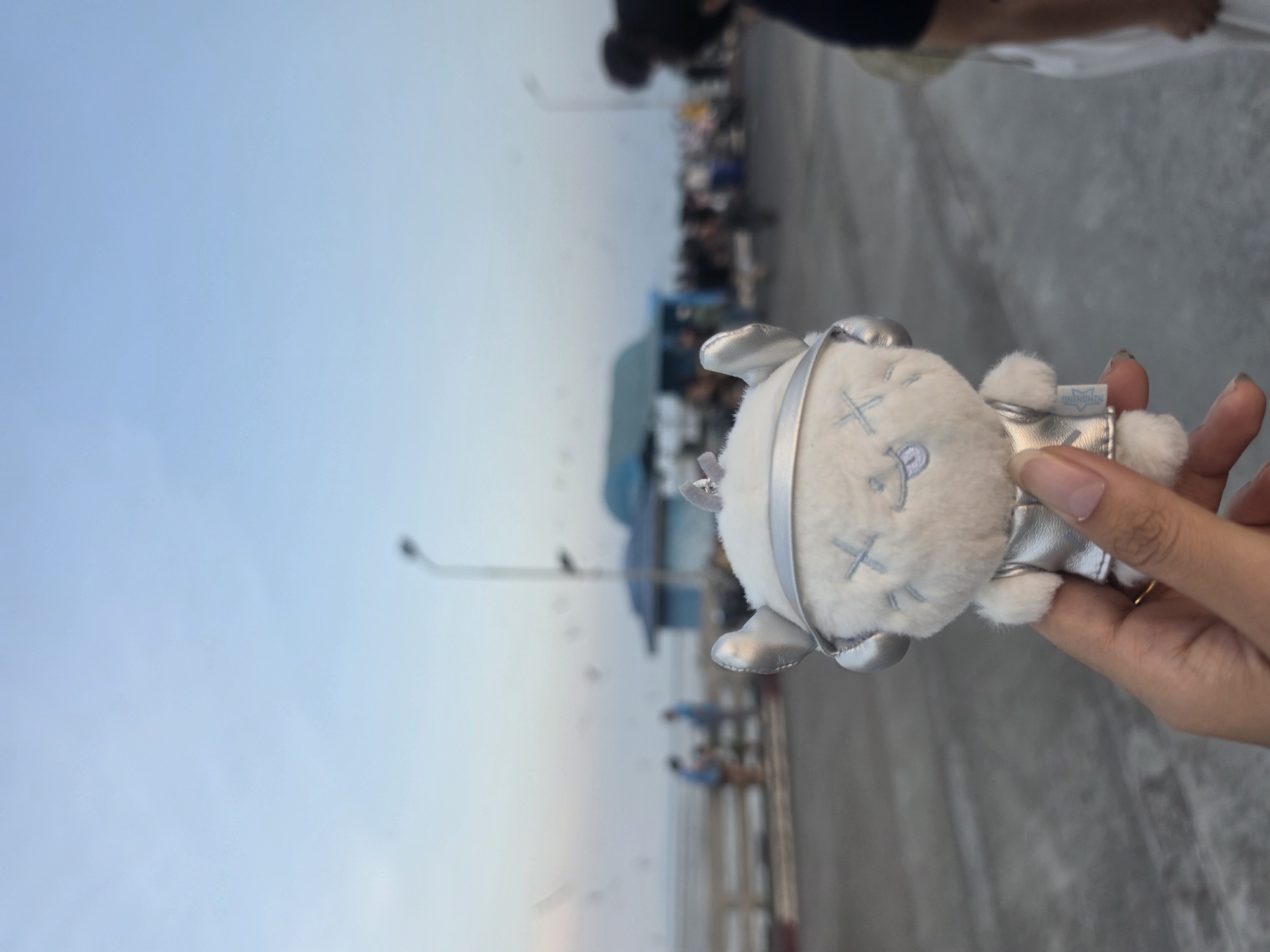 A hand holds a plush toy with a round, white, fluffy head and silver ears and limbs, against a background of a seaside location. The toy's face is embroidered with light blue "X" eyes and a curved mouth. The hand has painted fingernails and wears a gold ring.
The background shows a hazy sky meeting the horizon, with a pier or walkway extending into the distance. People are visible along the pier, but their details are indistinct due to distance and focus. Lamp posts line the walkway. The location is Bang Pu Subdistrict Municipality, Thailand.