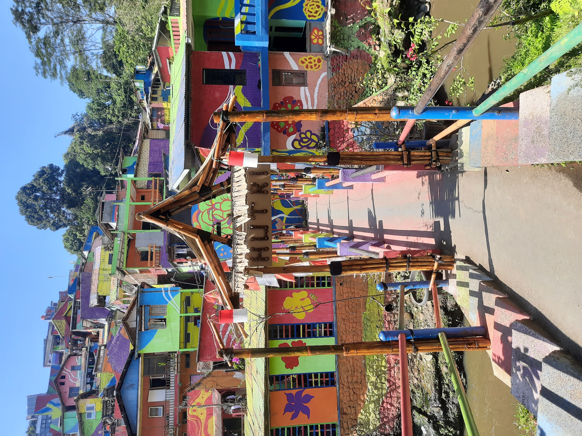 This is a photograph of a village built on a hillside, with houses painted in a vibrant array of colors. The village is located in Bandung, Indonesia, specifically in the Kampung Pelangi, known for its colorful architecture. In the foreground, a bridge constructed from bamboo and painted wood spans a body of water. The bridge has railings and steps that are also painted in bright colors, with shadows cast across them. Above the bridge, the houses climb up the hill, each facade adorned with different colors, patterns, and murals. Trees and vegetation are visible on the hillside, interspersed with the colorful buildings. The sky is clear and blue. There are no people clearly visible in the image, but laundry can be seen hanging from some of the houses. The overall scene depicts a lively and visually striking urban landscape.