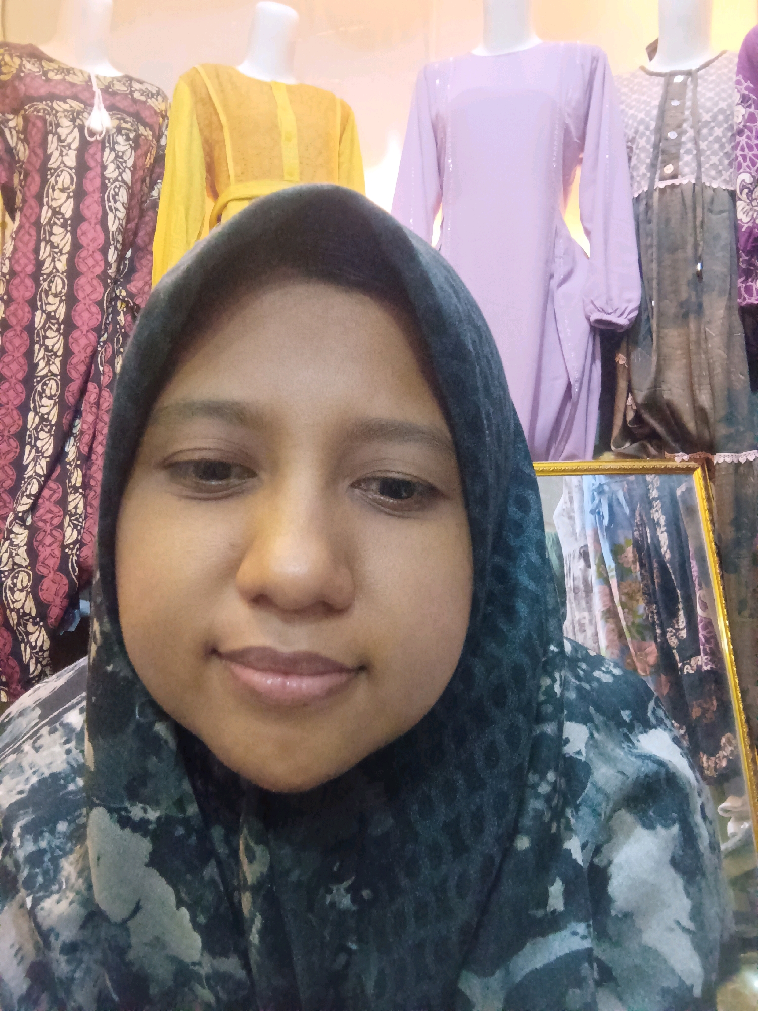 A close-up, front-facing selfie shows a young woman wearing a dark hijab with a subtle tie-dye pattern and a matching patterned garment. She is positioned in front of mannequins displaying various clothing items. To her left are mannequins in a dark patterned garment with intricate designs in red and cream, and a bright yellow garment. To her right, a mannequin wears a light purple long-sleeved dress with subtle embellishments, and another mannequin is dressed in a dark gray garment with white lace detailing. The background also shows a framed mirror reflecting more garments, some with floral patterns. The lighting is warm and slightly diffused. The scene appears to be within a clothing store, potentially in the city of Sumuragung, Indonesia.