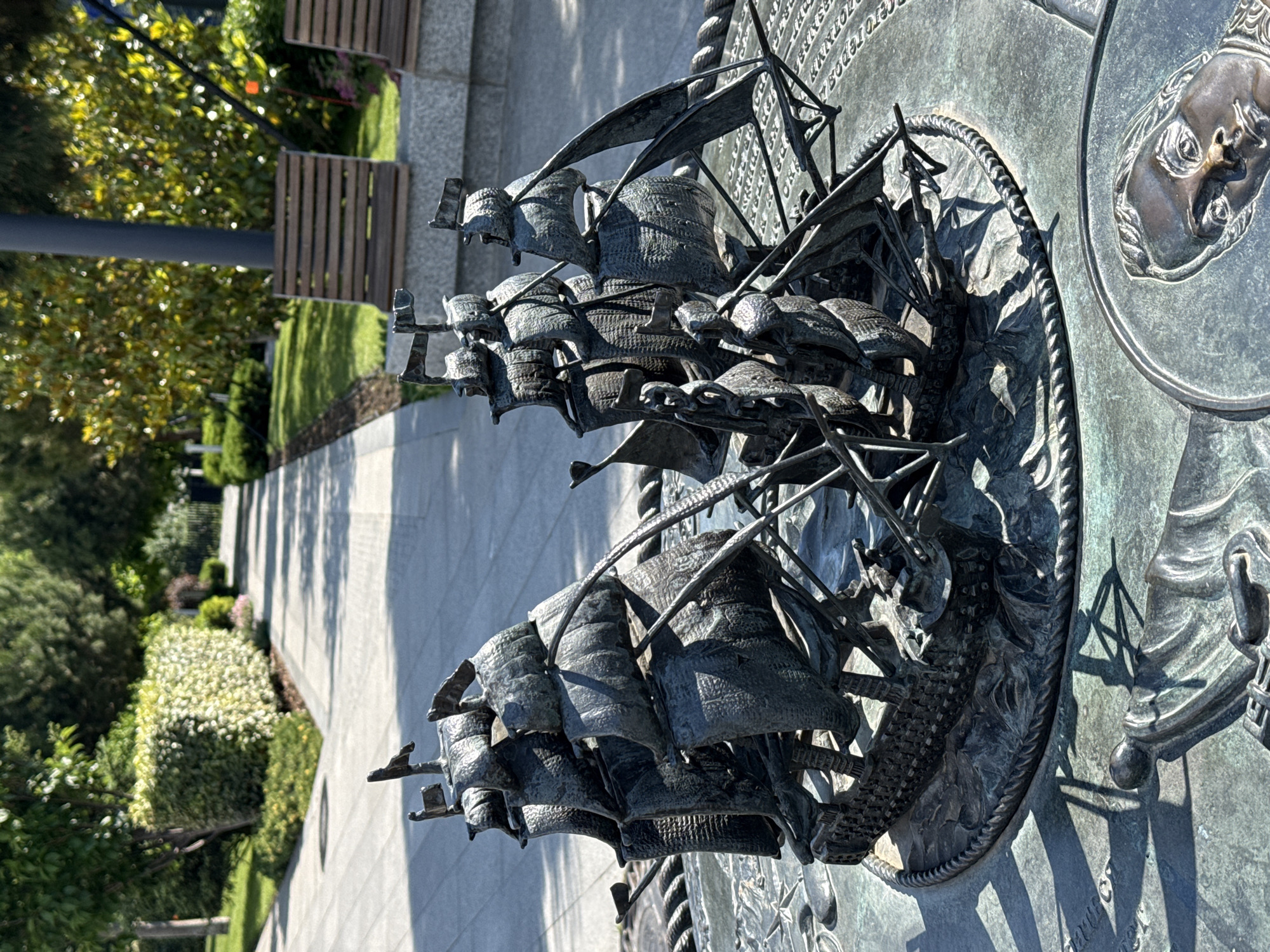 This is a close-up, eye-level shot of a bronze sculpture depicting a naval battle between several tall ships. The sculpture is placed on a round, textured pedestal, which has engraved text and a relief portrait of a man's face on its lower right side. The ships are intricately detailed with sails, masts, and rigging, appearing to be engaged in combat with cannon fire suggested by the dynamic arrangement. The foreground is dominated by the sculpture and pedestal, casting shadows on the metallic surface. In the background, a paved pathway lined with manicured bushes and trees creates a park-like setting. Wooden benches are visible further back along the path. The lighting suggests it is a sunny day.