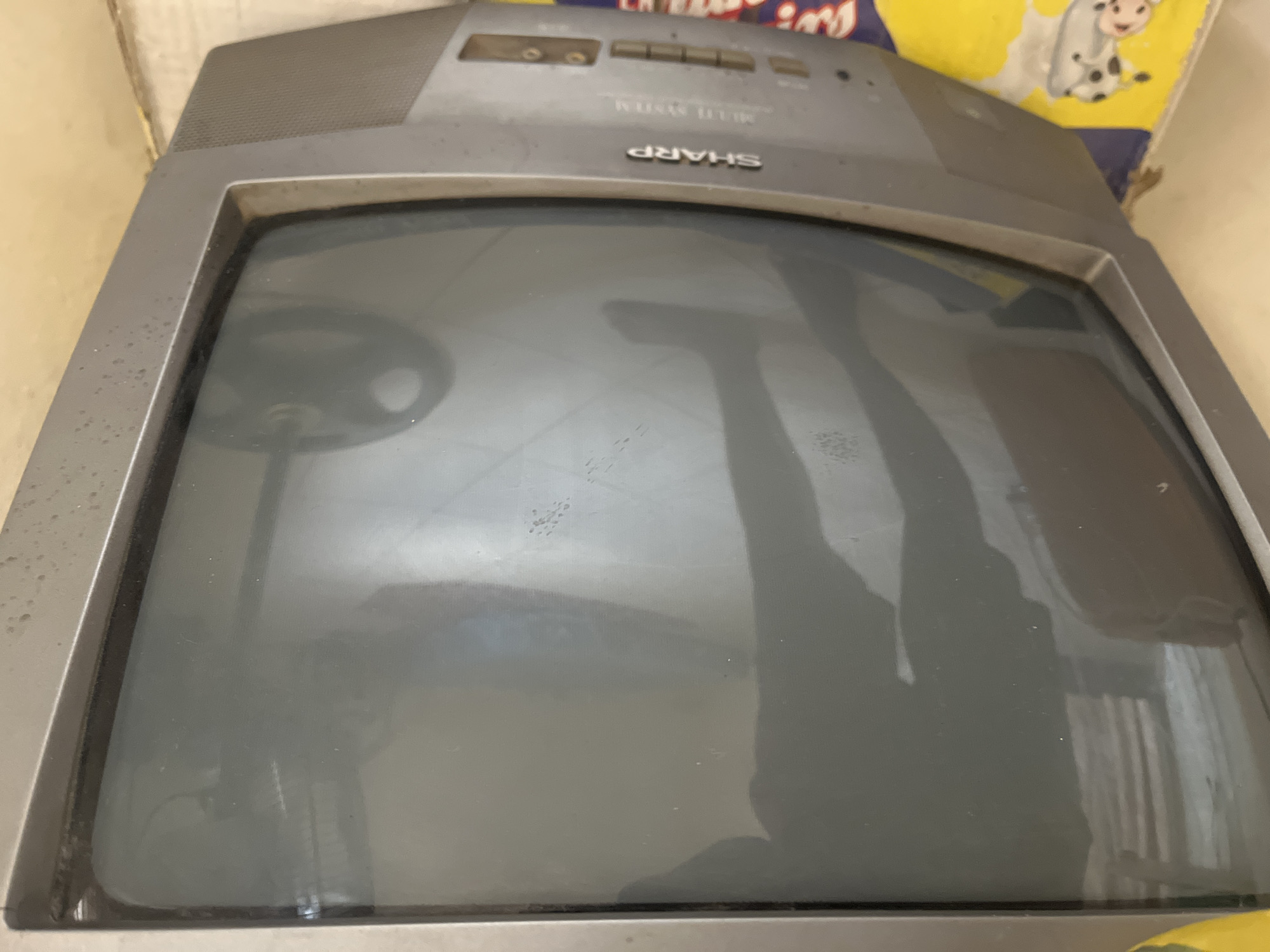A close-up shot shows the screen of an old-fashioned television set. The screen is dark, reflecting a shadowy figure of a person and a stand with a circular base. The television is gray and has a "SHARP MULTI SYSTEM" logo on its lower bezel. The set appears dusty and is placed on a colorful mat featuring cartoon cows. The scene is likely indoors, suggesting a domestic setting. The question of location in Jalingo, Nigeria, cannot be determined from the visual information.