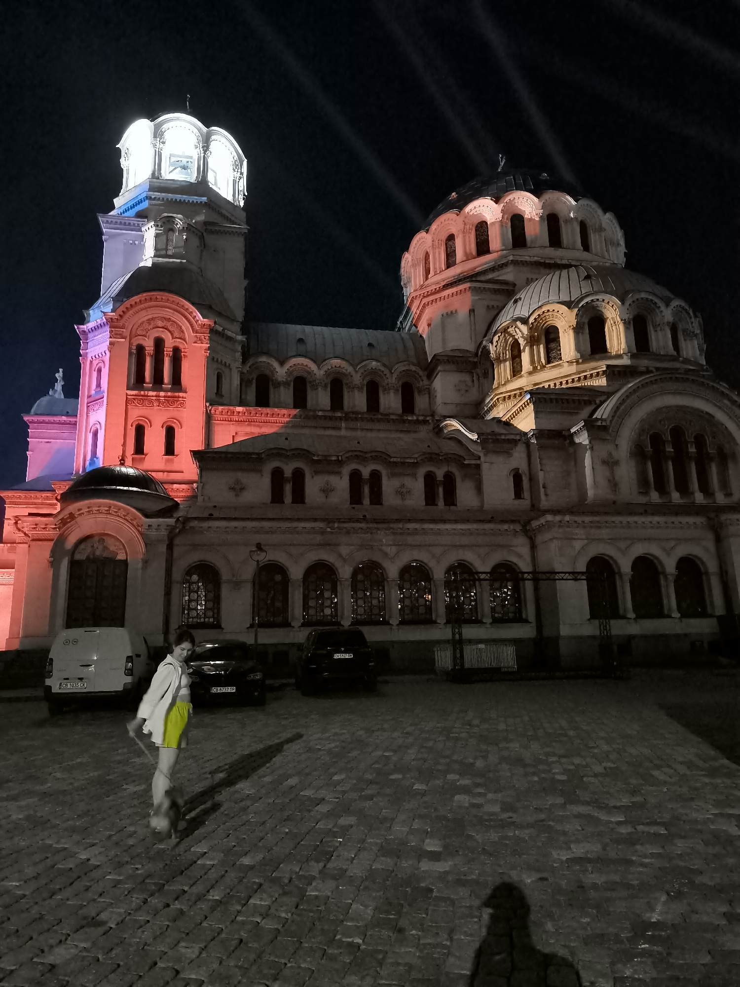 An eye-level, full-shot photograph captures a woman and her dog on a cobblestone plaza in front of the St. Alexander Nevsky Cathedral in Sofia, Bulgaria, at night. The cathedral's exterior is illuminated with vibrant red and yellow lights. The woman, wearing a white unbuttoned shirt over a white top and bright yellow shorts, is holding a leash attached to her small, light-colored dog. Both are casting long shadows on the plaza. The woman appears to be looking down at her dog. In the background, several vehicles are parked, including a white van and two dark sedans. The sky above is dark, with beams of light emanating from the cathedral's towers. The overall atmosphere is one of quiet nighttime activity in a prominent urban setting.