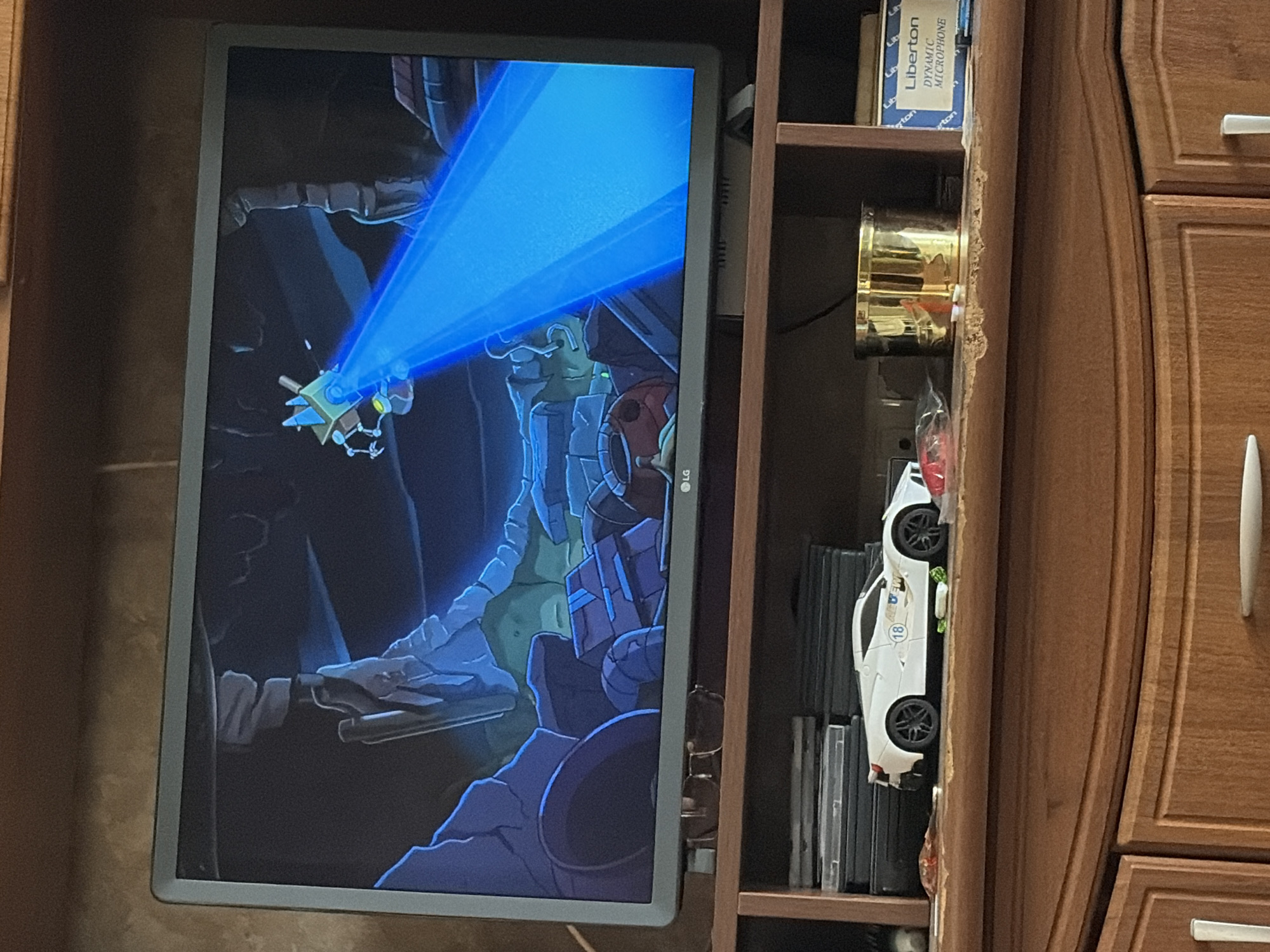 The image shows a television screen displaying an animated scene of a small, yellow, robotic creature with blue triangular accents on its head, shooting a bright blue beam of light forward. The background of the animated scene appears to be a rocky, cavernous environment with blue and purple hues. Below the television, on a wooden shelf, sits a white toy sports car with the number "18" on its side and some decals. To the right of the car is a stack of what appear to be CDs or DVDs, and further to the right is a brass cylindrical object and a box labeled "Liberton DYNAMIC MICROPHONE". The television is resting on a wooden cabinet with two visible drawers, one of which has a silver handle. The overall scene is indoors, likely a living room or entertainment area.