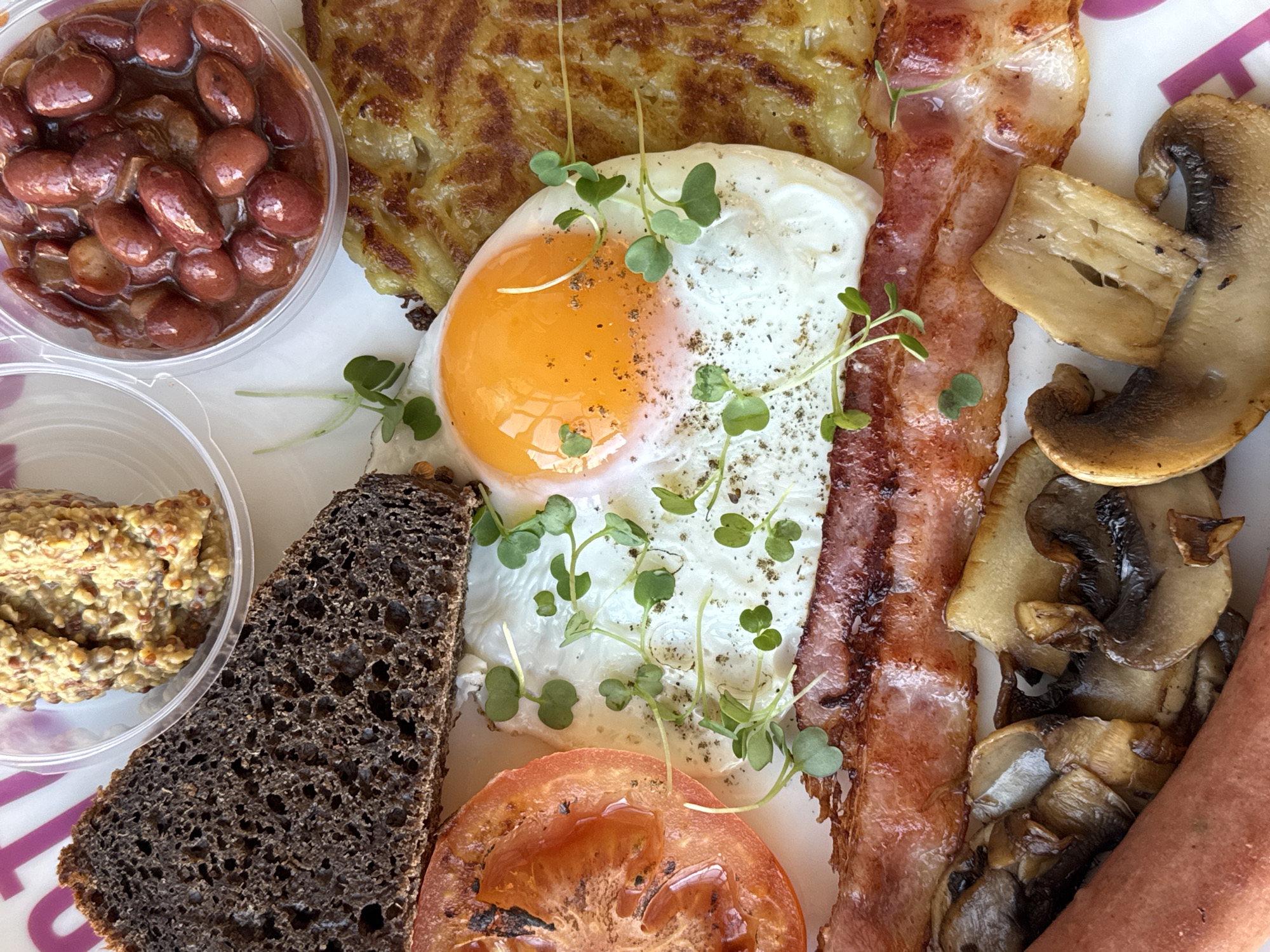 A high-angle, close-up shot presents a fully-loaded breakfast plate, possibly in Saint Petersburg, Russia, based on the availability of such plates. The components of the meal include a clear container filled with beans, a crispy potato pancake (rösti), a sunny-side-up egg garnished with microgreens, two slices of bacon, sautéed mushrooms, a sausage, a slice of grilled tomato, a slice of dark rye bread, and a clear container of coarse-grain mustard. The meal components are arranged in close proximity on a white plate. The plate sits on top of a patterned pink surface. No people are visible.