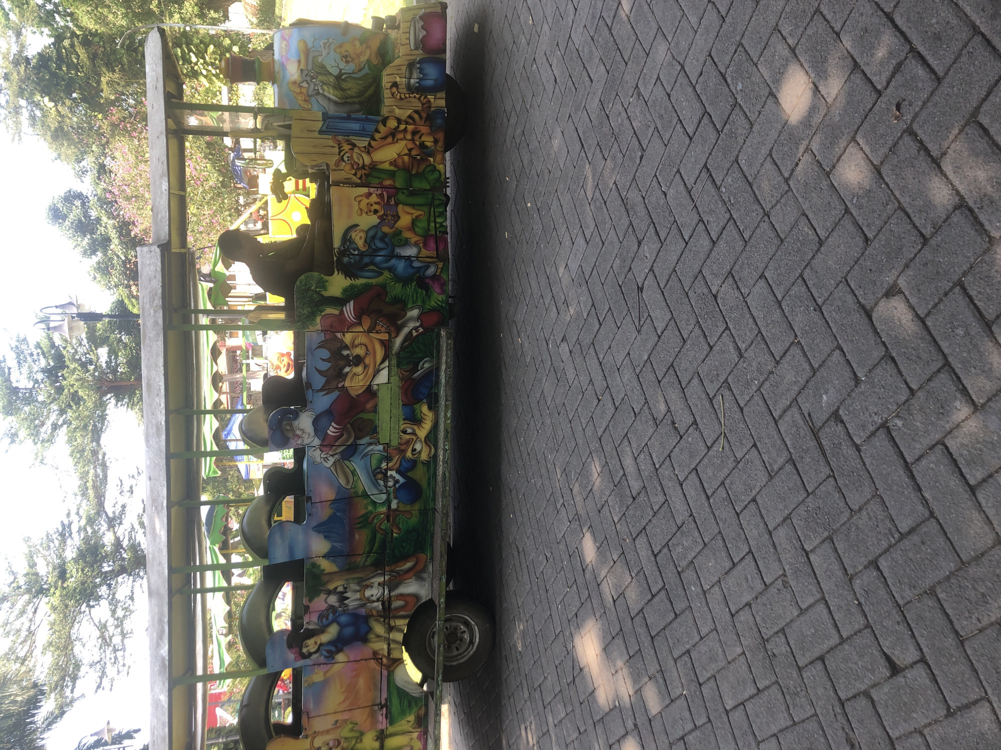 A colorful amusement park train is parked on a brick-paved pathway outdoors. The train cars are decorated with cartoon characters, including Snow White, Winnie the Pooh, Tigger, Eeyore, and the Tasmanian Devil. A person, possibly the driver, is seated in the front car of the train. The background shows trees and other park attractions, suggesting the location is within a park. The image was taken on a sunny day. The specific location in the city of Pasuruan, Indonesia, is identified as Taman Safari Indonesia II, specifically at the Mini Train area.