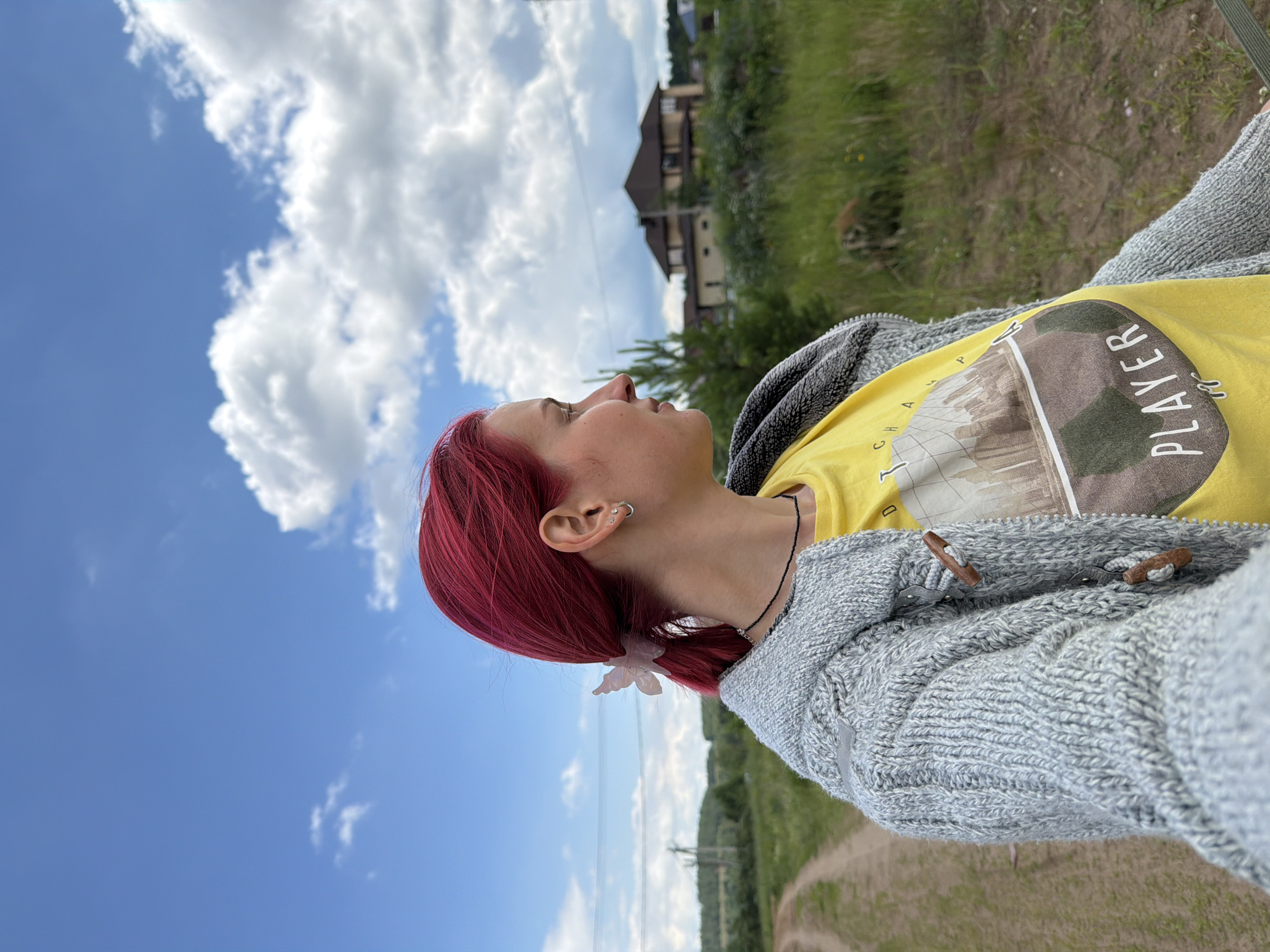 A young woman with vibrant pink hair tied back with a butterfly clip stands outdoors. She is wearing a bright yellow t-shirt with a graphic print and a light gray knitted cardigan. The woman is looking to her right, with a profile view of her face. The background features a dirt road leading into a grassy field dotted with trees and bushes. In the distance, several houses with brown roofs are visible. The sky above is a bright blue with fluffy white clouds. This scene appears to be in a rural or suburban area, possibly on the outskirts of Chaikovsky, Russia.