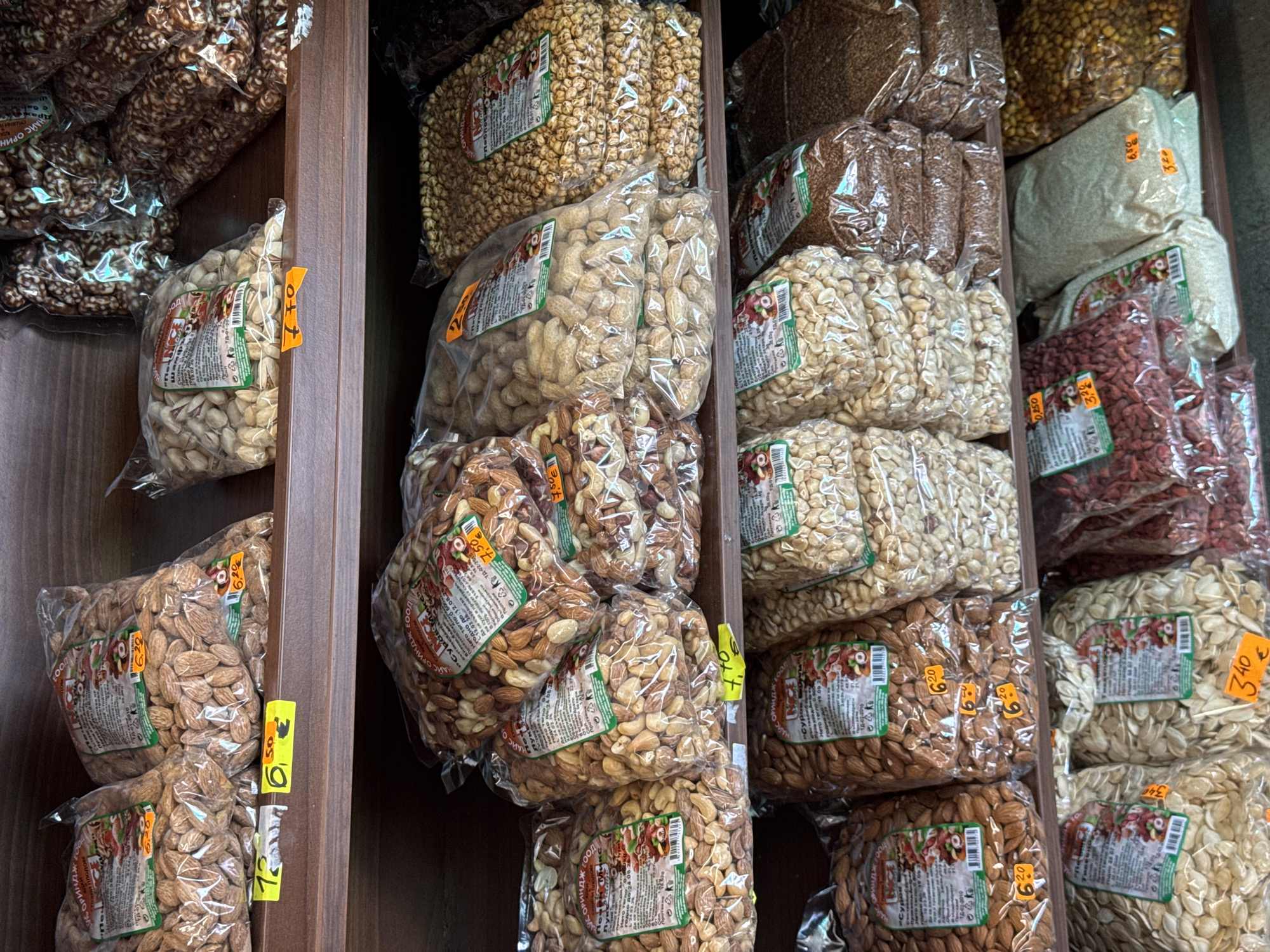 A close-up shot depicts shelves filled with plastic bags of various nuts and seeds, likely in a market or store in Burgas, Bulgaria. The bags are neatly stacked and each has a product label with a depiction of a red squirrel and a price tag in Euros. The nuts include pistachios, peanuts, almonds, brown flax seeds, chick peas, goji berries, pumpkin seeds. The shelving is made of dark wood, and the overall lighting in the scene suggests an indoor setting.