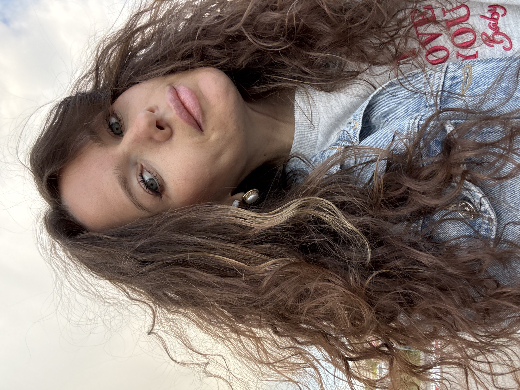 A tilted close-up shot features a woman with long, curly brown hair that frames her face and cascades over her shoulders. She has fair skin, green eyes, and pink lips, and is wearing a denim jacket over a grey sweatshirt. Part of the text on the sweatshirt is visible, reading "Baby I love You". The background is indistinct but appears to be a sky with light cloud cover and greenery. The location is not definitively identifiable as Moscow, Russia without further information.