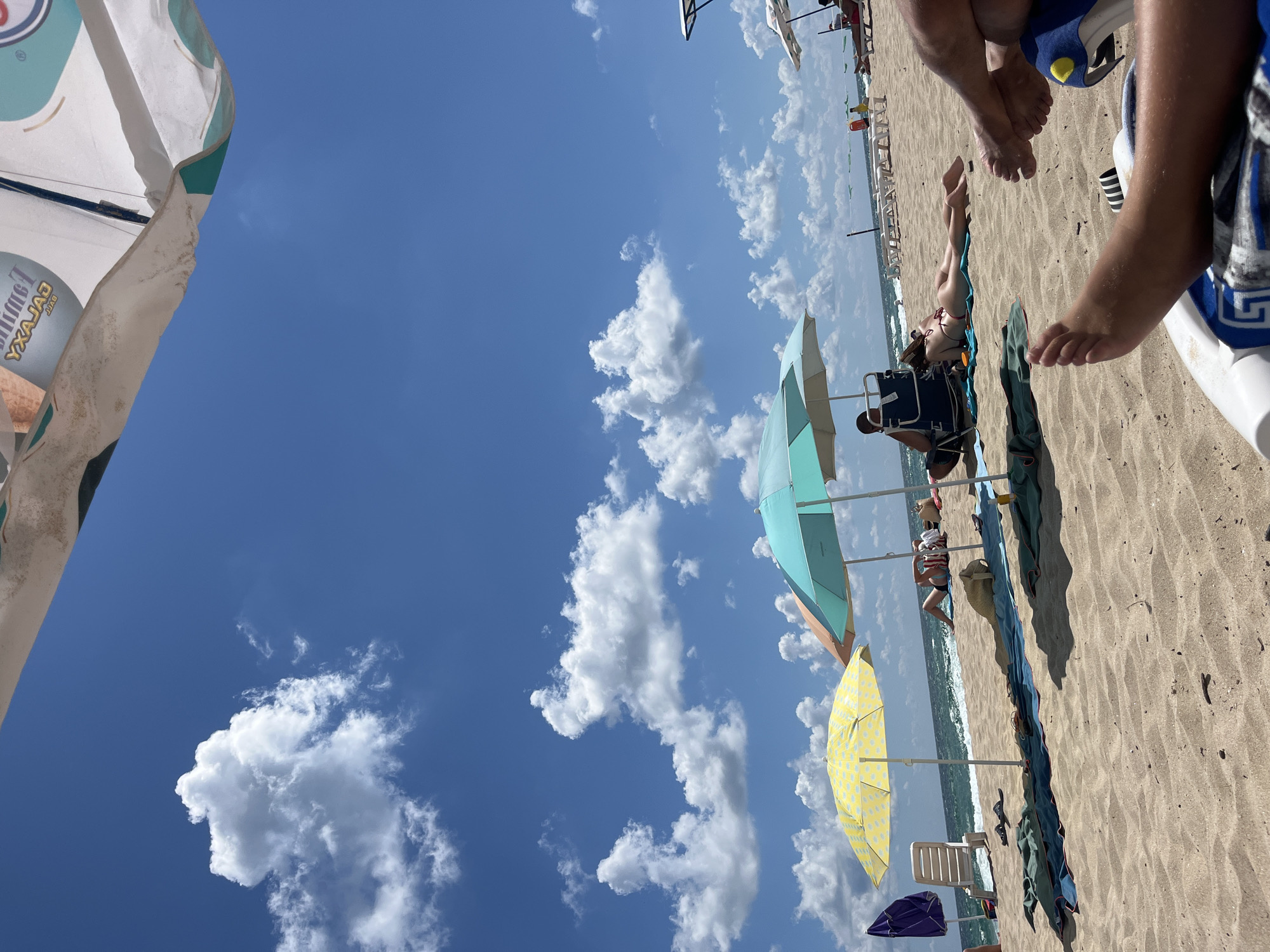 The image depicts a sunny day at a beach in Primorsko, Bulgaria. In the foreground, the sandy beach is visible with a person's legs and feet in the lower right, resting on a blue and white lounge chair. Further back, several beach umbrellas provide shade. A yellow umbrella with a polka dot pattern is prominent, and next to it is a teal and white umbrella. People are seen relaxing under the umbrellas and on lounge chairs. A person is sunbathing on a blue towel under the teal umbrella. In the background, the sea with moderate waves is visible under a bright blue sky dotted with fluffy white clouds. Some lounge chairs are set up further back on the beach. A purple umbrella is visible on the left side of the image. The overall scene conveys a typical summer day at the beach.