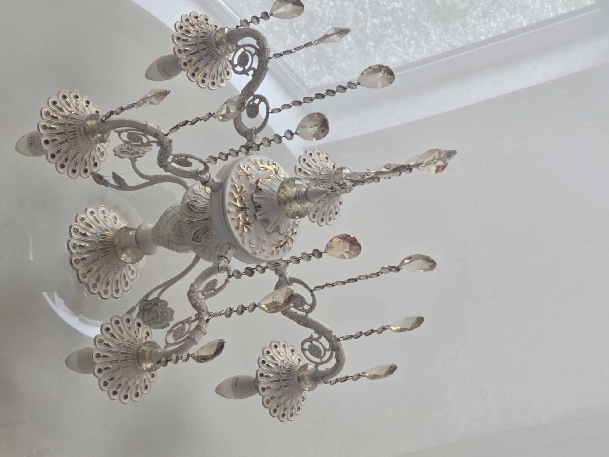 The image shows a white chandelier with gold accents hanging from a white ceiling. The chandelier has multiple arms, each adorned with decorative floral elements and crystal teardrop pendants. Light bulbs are visible at the end of some arms. The overall aesthetic is ornate and classic. The location is a room in Avtury, Russia, with a white wall and a sheer white curtain visible in the background, suggesting a room with natural light. There are no people or animals present in the image.