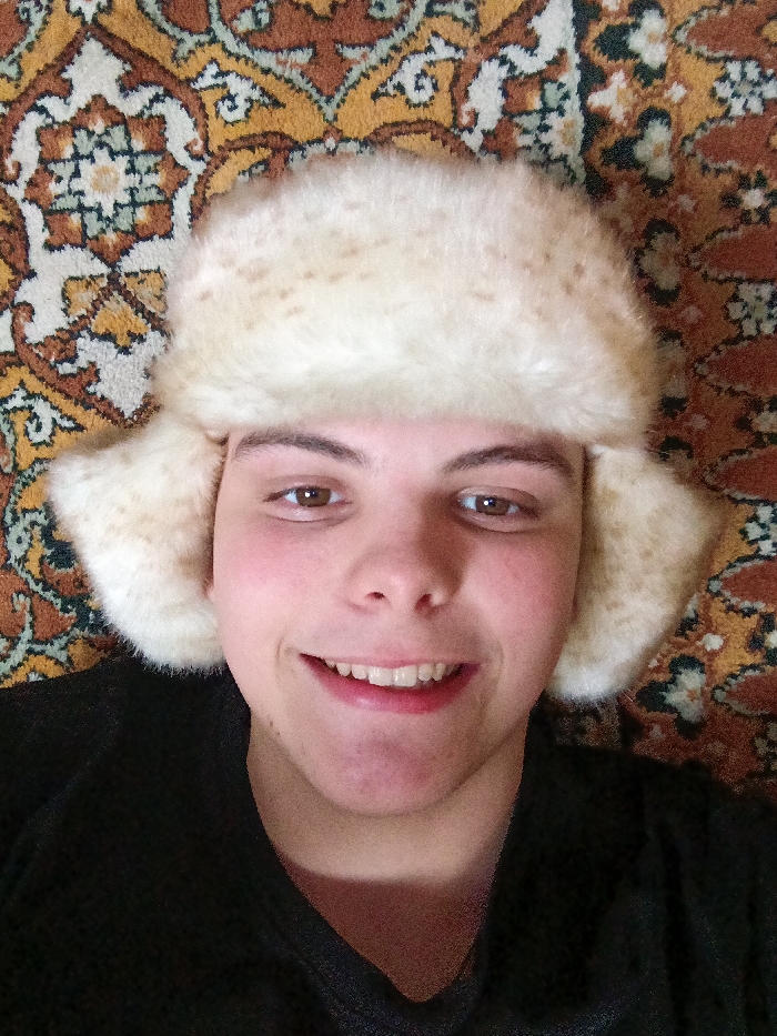 A smiling person with light skin, brown eyes, and short dark hair, wearing a furry white hat with ear flaps, is positioned against an intricately patterned rug of brown, orange, and white hues. The individual is dressed in a dark shirt. The close-up shot highlights the person's facial features and the unique texture of the fur hat, creating a contrast against the busy background of the rug.