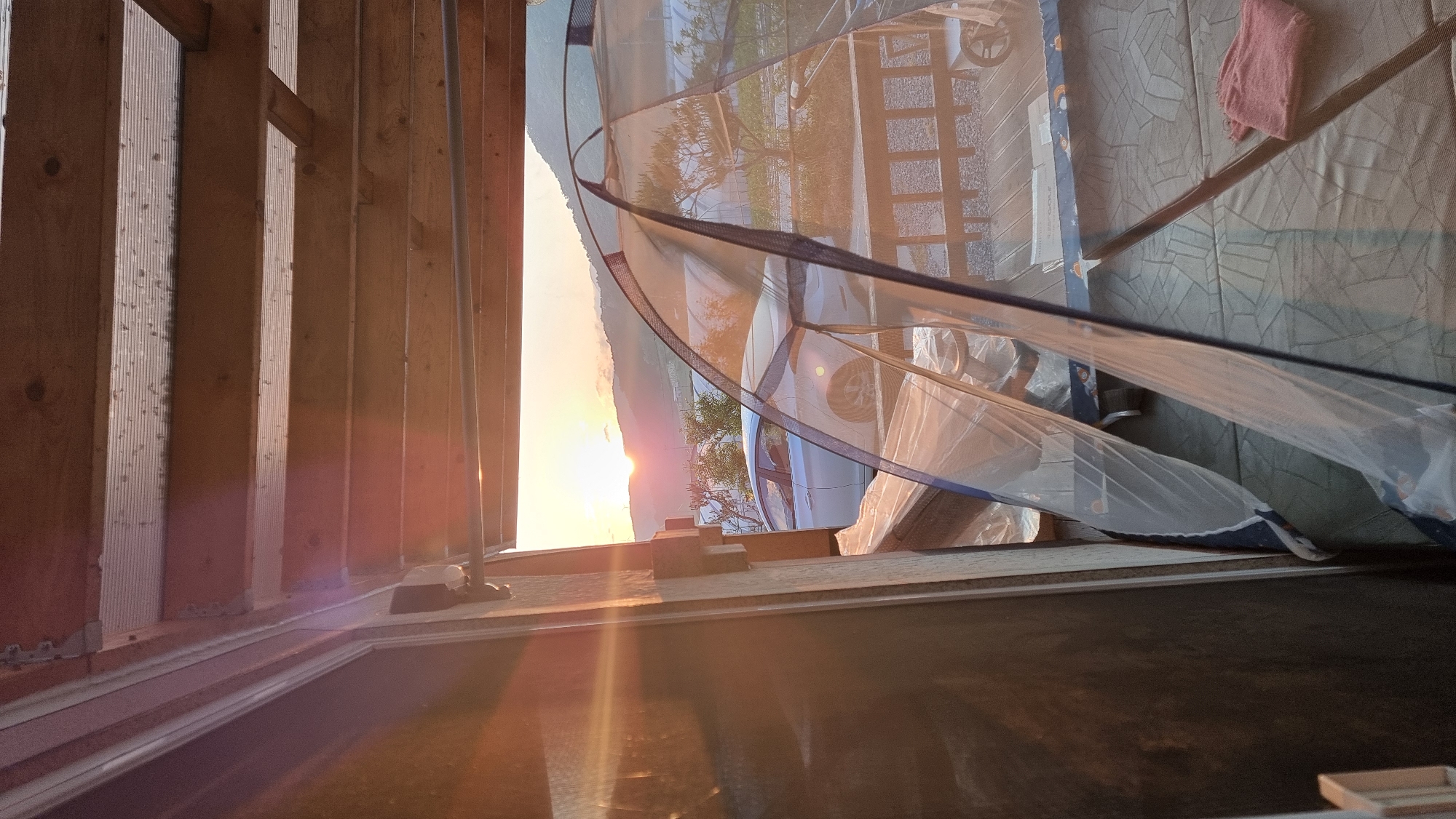 The image depicts a scene in Dunnae-myeon, South Korea, capturing the interplay of interior construction and an outdoor view obscured by a screen. In the foreground, unfinished wooden studs, characteristic of a construction project, run vertically along the left side of the frame. Light from the setting sun streams between the studs, casting an orange glow.
The background features a partially obstructed view through a window or opening covered with a white netting or screen with blue trim. Behind the screen, a car is visible, parked in front of what appears to be a house and a street. The setting sun casts a silhouette of the distant hills and trees. A pink towel lies on the other side of the screen.