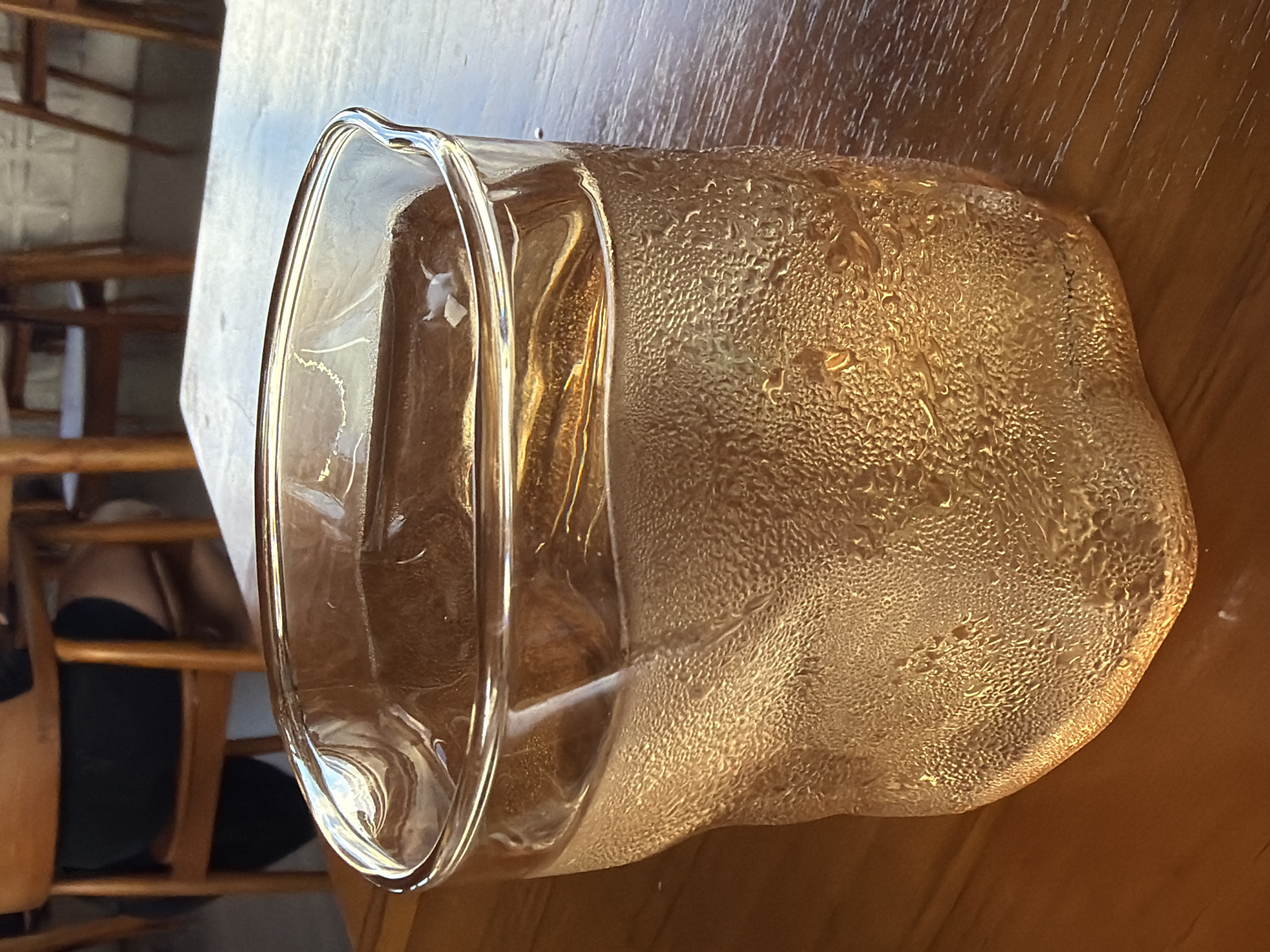 The image shows a glass on a wooden table. The glass is slightly tilted to the left. The glass has condensation on the outside. Some wooden chairs are visible in the background. The photo was taken in Kuta Selatan, Indonesia.