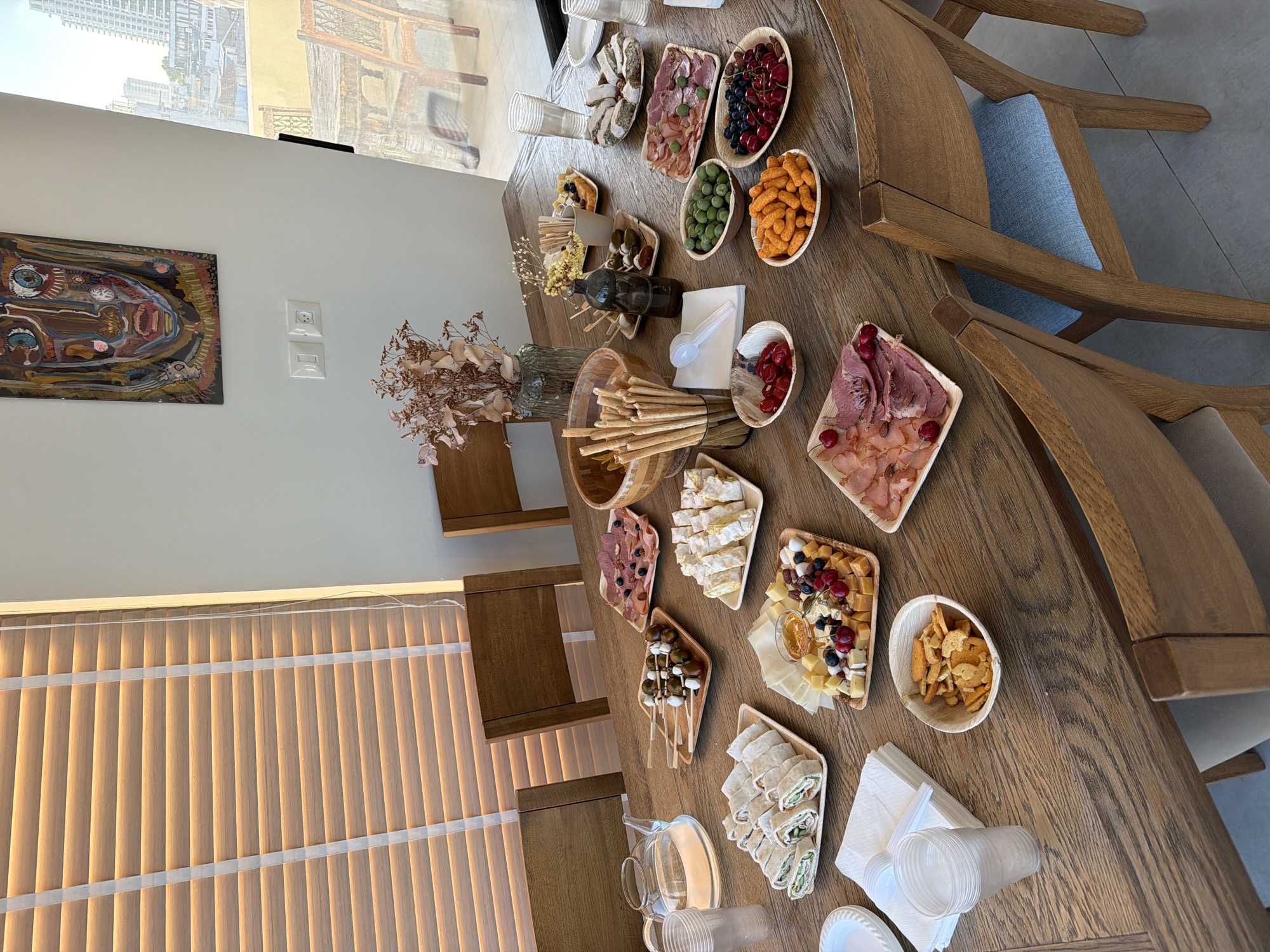 A horizontal, high-angle, eye-level shot captures a large, rustic wooden table laden with an assortment of appetizers. The table is set for a gathering, with several wooden chairs around it. In the background, a large window reveals a view of Tel Aviv, Israel, with buildings and a cityscape under a bright sky. The room features a modern interior with minimalist decor.
The table is covered with various platters of food, including sliced meats, cheeses, olives, berries, and rolled wraps. Bowls of snacks like crackers and cheese puffs are also present. A glass teapot with water sits on the left side of the table, along with plastic cups and napkins. A decorative dried flower arrangement adds a touch of natural element to the scene. The overall impression is one of a sophisticated and well-catered event.