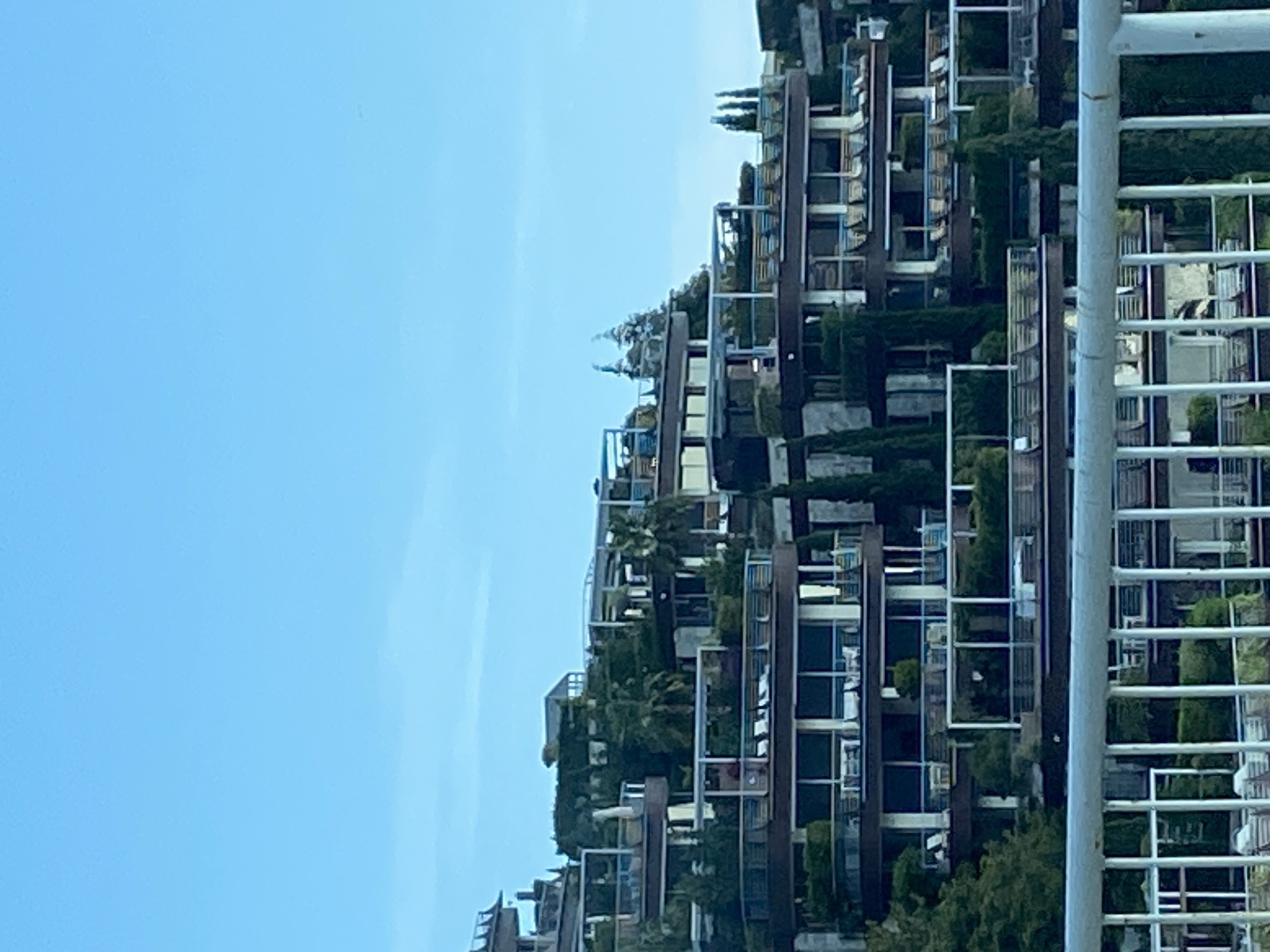 The image depicts a modern residential building complex against a clear blue sky. The building features a tiered structure with balconies and vertical gardens, integrating greenery into the facade. The architecture is angular and contemporary, with repeating geometric elements. A white fence or railing is visible in the foreground, slightly obscuring the lower right portion of the building. The location is likely Budva, Montenegro, given the style of architecture and the presence of dense vegetation. The image presents an urban environment where nature and architecture are blended.