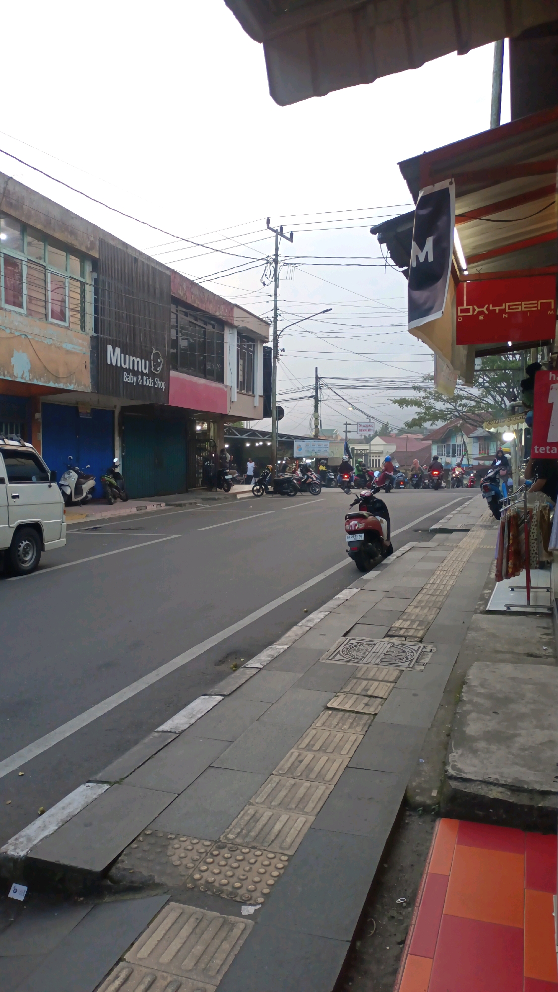 The image captures a street scene in Padang Panjang, Indonesia, likely during the day given the ambient light. The perspective is from a sidewalk looking towards a street lined with buildings and a variety of parked and moving motorcycles.
On the left side of the street, a white van is parked near a building with storefronts. One storefront displays a sign that reads "Mumu Baby & Kids Shop". The building itself appears to have multiple windows and a textured facade. Further down the street, more shops and buildings are visible, some with awnings.
The street is paved and marked with a white lane divider. Numerous motorcycles are parked along the side of the road and others are in motion, indicating a bustling commercial area. Some individuals are visible near the shops and on the motorcycles.
The sidewalk in the foreground is made of concrete tiles and includes tactile paving for the visually impaired. There are also some manhole covers visible on the sidewalk. To the right of the sidewalk, more shops are present, with clothing racks displaying apparel outside one establishment. A red and orange tiled area is also visible at the edge of the sidewalk.
Overhead, power lines and poles are prominent, common in urban Indonesian streetscapes. The sky is overcast and bright, suggesting subdued natural lighting. The overall impression is a typical urban street environment in Indonesia, with a focus on commerce and transportation.