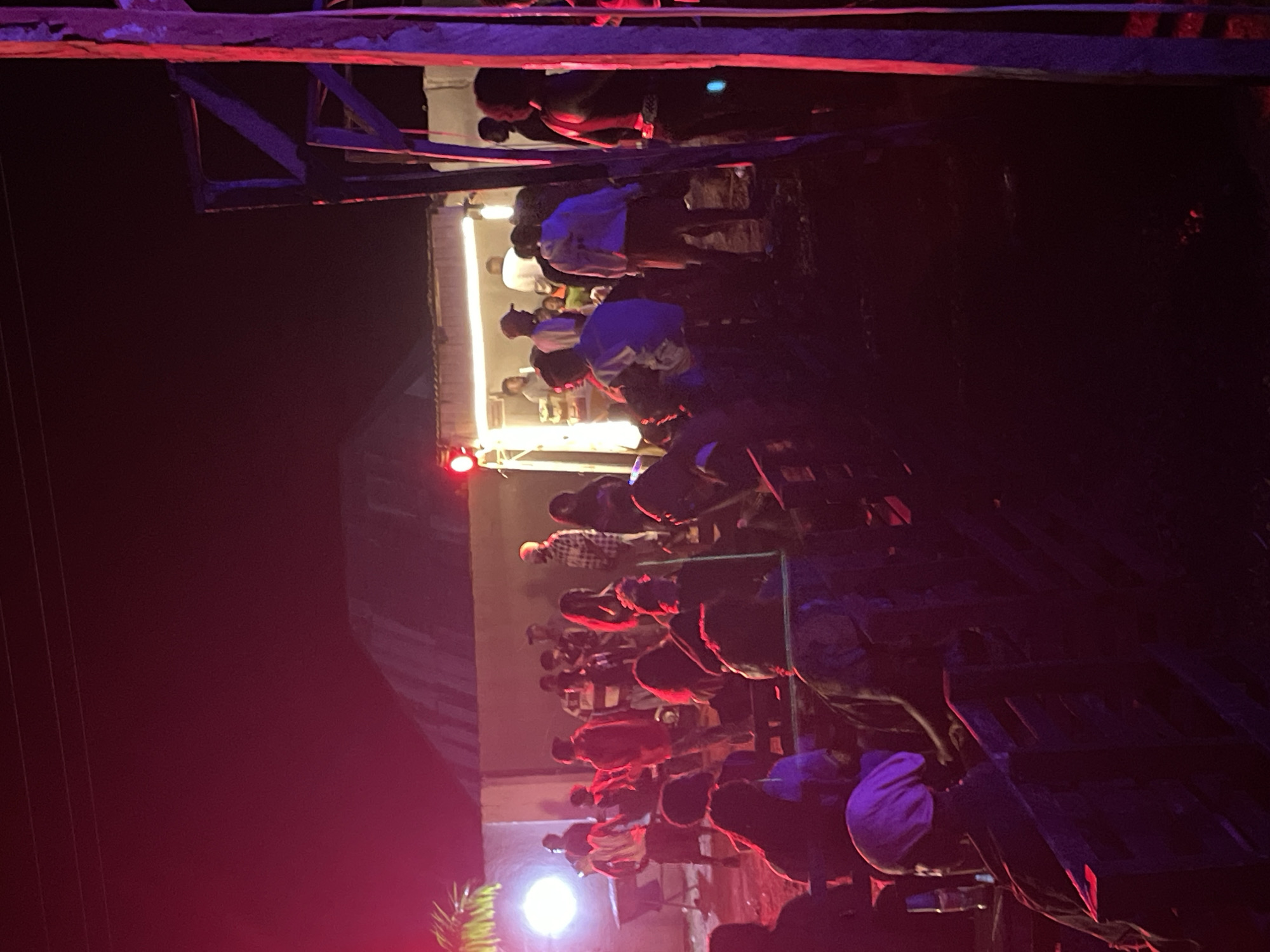 The image depicts an outdoor gathering at night, possibly in Durumi, Nigeria, with red and purple lighting illuminating the scene. A group of people are visible, some standing, others seated on wooden structures resembling benches. In the background, a building with a corrugated roof is present, and a food stall or small structure with a light source above it. A wooden scaffolding or frame is visible on the right side of the image. The overall atmosphere appears to be that of a social event or a small festival.