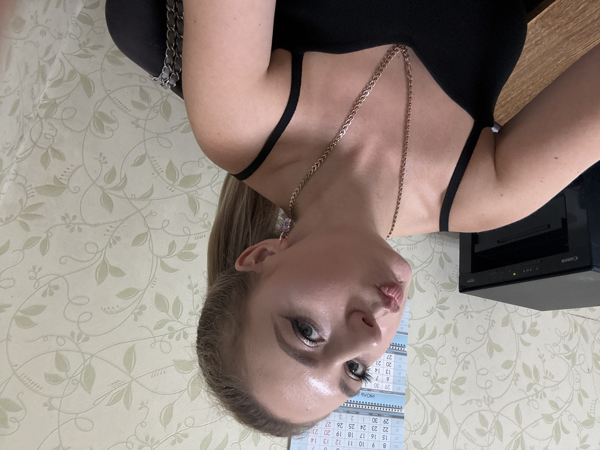 A woman with blonde hair in a ponytail makes a kissing face at the camera, her head tilted to the left. She is wearing a black tank top and a gold chain necklace. A printer is visible to her left. A calendar with Cyrillic text and numbers is on the wall behind her. The wallpaper is a light color with a subtle floral pattern. The scene appears to be indoors, possibly in an office or home setting. The image quality is somewhat grainy and overexposed in places, particularly on the woman's forehead and cheeks.