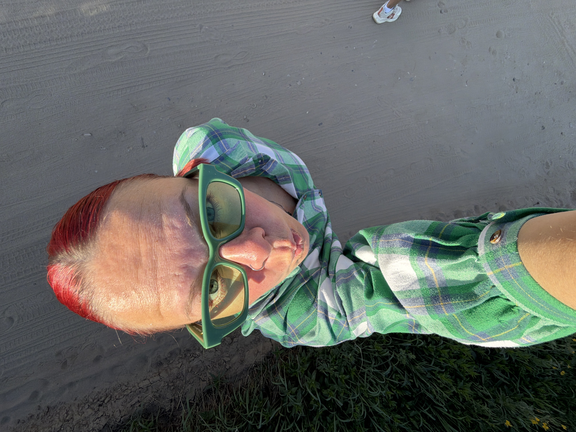 A high-angle shot shows a person with red hair, green-framed sunglasses, and a green plaid shirt. The individual appears to be standing on a surface comprised of dirt, sand, and grass. A shoe is visible in the background. The precise location within Chaikovsky, Russia cannot be determined from the available information.