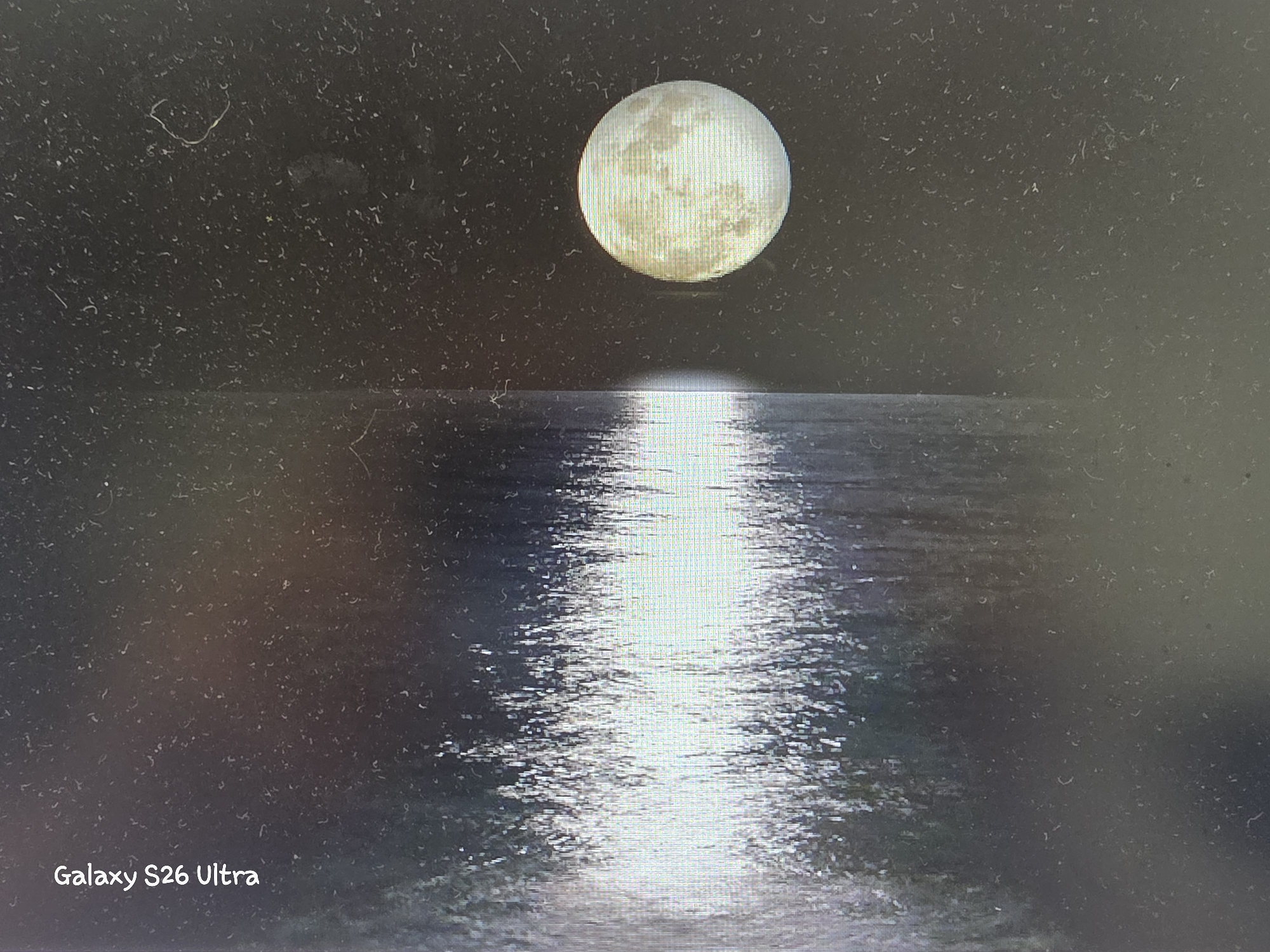 The image shows a full moon reflected on the surface of the water. The moon is a bright, luminous orb in a dark, starry sky. The reflection of the moon creates a shimmering pathway of light across the water. The scene is calm and serene, evoking a sense of peace and tranquility. The image appears to be taken at night, possibly during a clear and cloudless evening. The overall impression is one of natural beauty and the quiet majesty of the night sky. There is text visible in the bottom left corner that says "Galaxy S26 Ultra". The location context is Plovdiv, Bulgaria, however, there are no specific visual cues within the image to confirm or deny this location.
