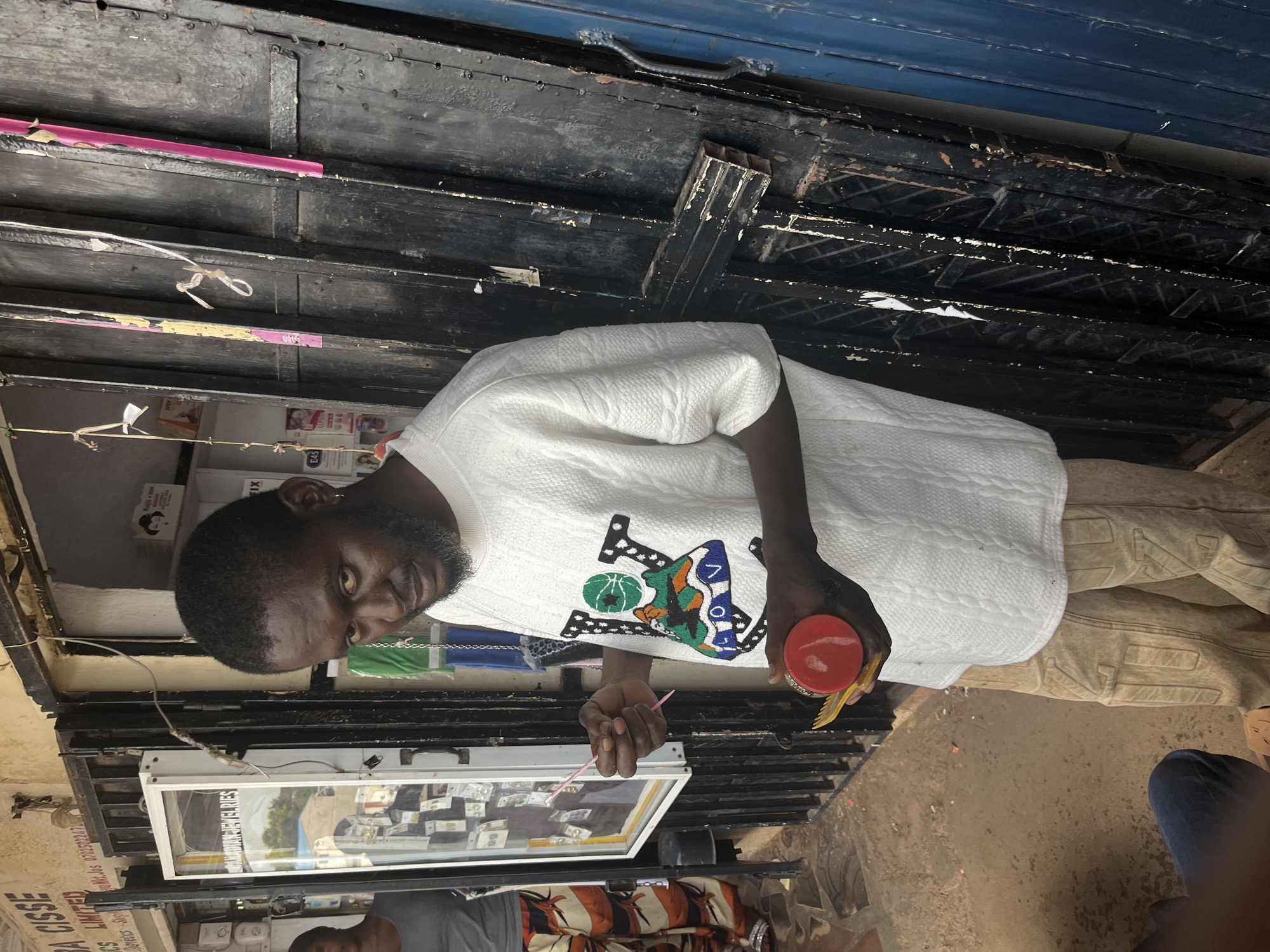 A man stands in front of a shop in Jos, Nigeria, holding a red circular container and a yellow comb. He is wearing a white textured t-shirt with a colorful graphic design and tan pants. To his left, a glass display case filled with jewelry is visible. Behind him, a dark metal door with peeling paint and a blue metal door are also present. The ground is covered in dirt and debris. The man is looking directly at the camera.