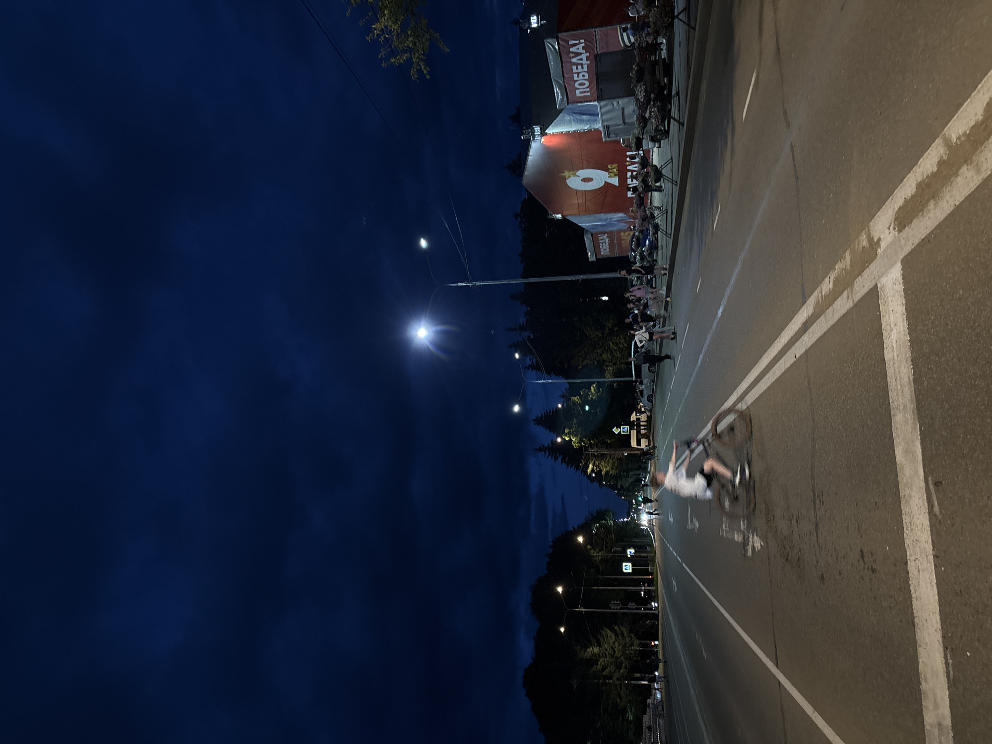 The image depicts a street scene at night in Birobidzhan, Russia. The sky is a deep, dark blue, suggesting late evening or night. There are several streetlights visible, illuminating the asphalt road marked with white lines.
The main subject in the foreground is a person on a bicycle, captured in mid-action, appearing to be riding or performing a stunt. The person is wearing a light-colored shirt and dark shorts. The bicycle is also visible, with its wheels in motion.
In the background, along the side of the street, there is a building or structure adorned with large banners. One prominent banner features a large red number "9" and the word "МАЯ" (May), alongside the word "ПОБЕДА!" (Victory!). These elements suggest a commemoration or celebration related to Victory Day, which is celebrated on May 9th in Russia. Other banners with the word "ПОБЕДА!" are also visible.
A crowd of people can be seen gathered along the street and in front of the building, some seated on chairs, watching the events. Trees and foliage line the far side of the road, adding to the urban setting. The overall atmosphere seems to be one of a public gathering and celebration at night.