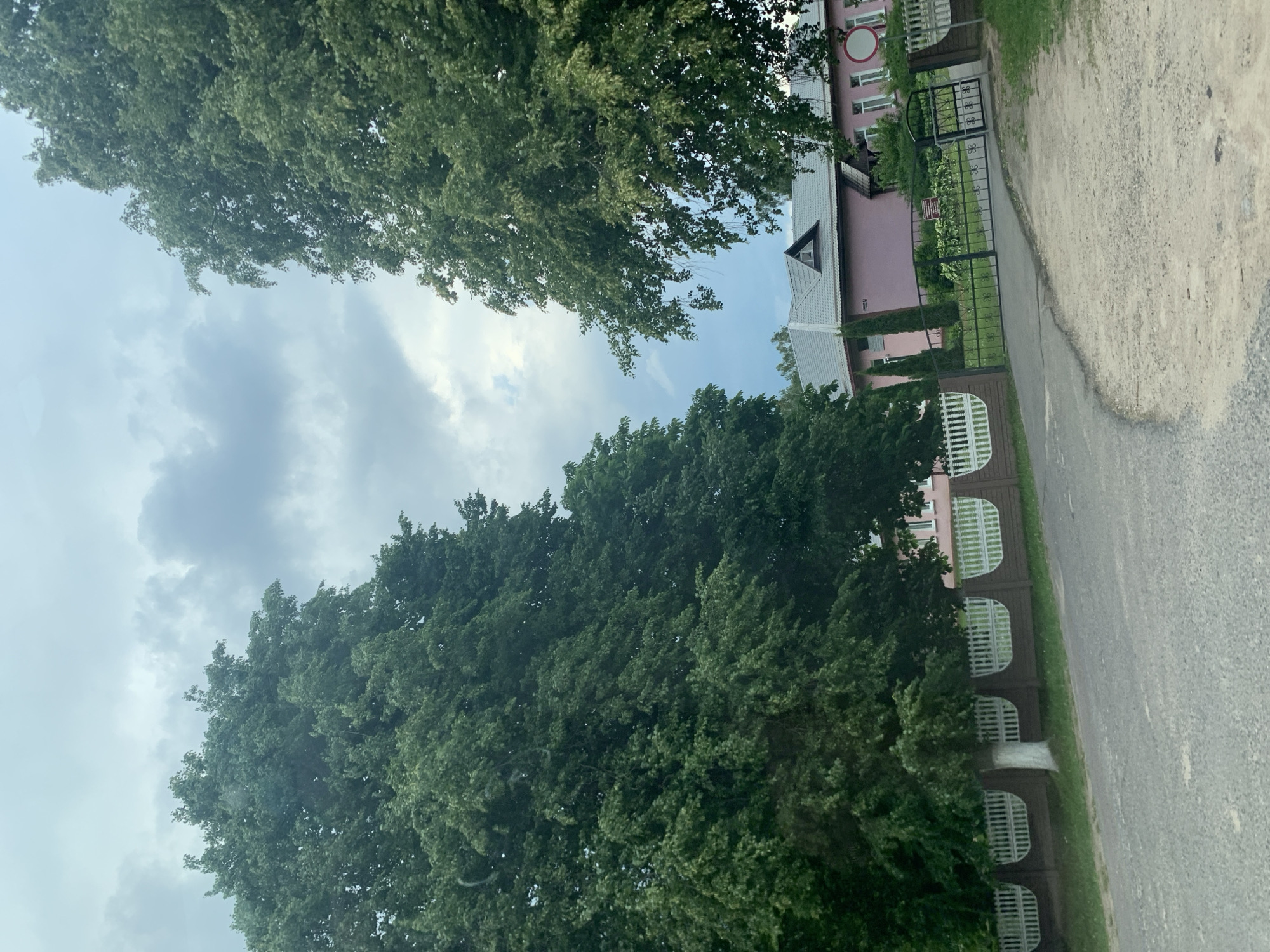 The image captures a street view in Слабада-Кучынскі сельскі Савет, Belarus. On the left and right sides of the frame, two large, mature trees with dense green foliage dominate the scene. Between them, a light pink building with a grey, gabled roof and a small triangular dormer window is visible. The building appears to be a residential or institutional structure, with several windows and a round, red and white sign on the right side. A decorative black metal gate with an ornate design provides an entrance to the property, flanked by a brown and white fence. A paved road, with a gravelly shoulder, leads towards the gate. The sky above is overcast with a mix of grey and white clouds.