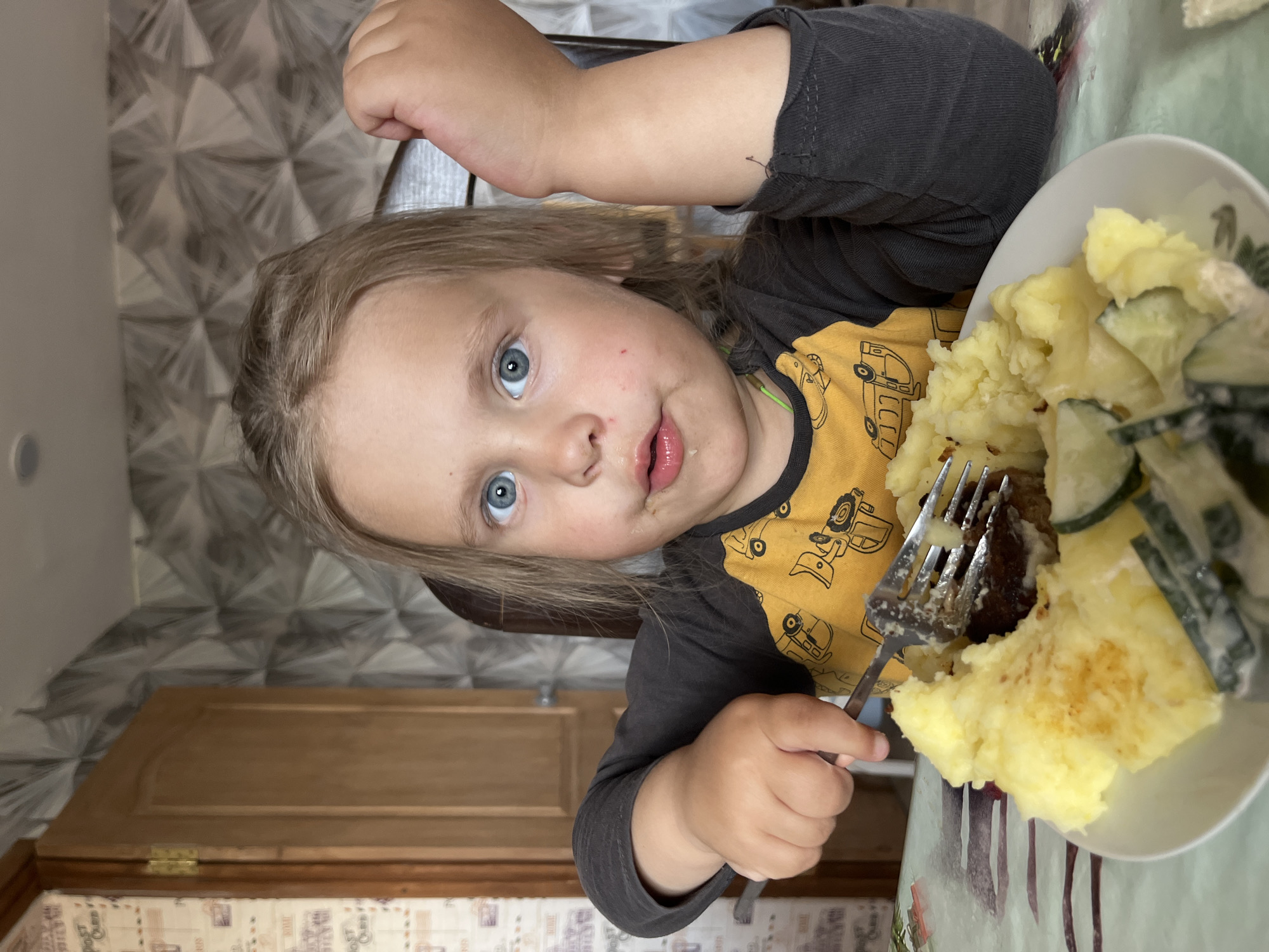 A young child with blonde hair and blue eyes is sitting at a table in Chaikovsky, Russia. The child is holding a fork with food on it, consisting of mashed potatoes, meat, and sliced cucumbers. The child wears a long-sleeved shirt with a yellow and grey design of construction vehicles. The table is covered with a patterned tablecloth, and there is a patterned wallpaper in the background. A wooden door is visible in the lower-left corner of the frame.