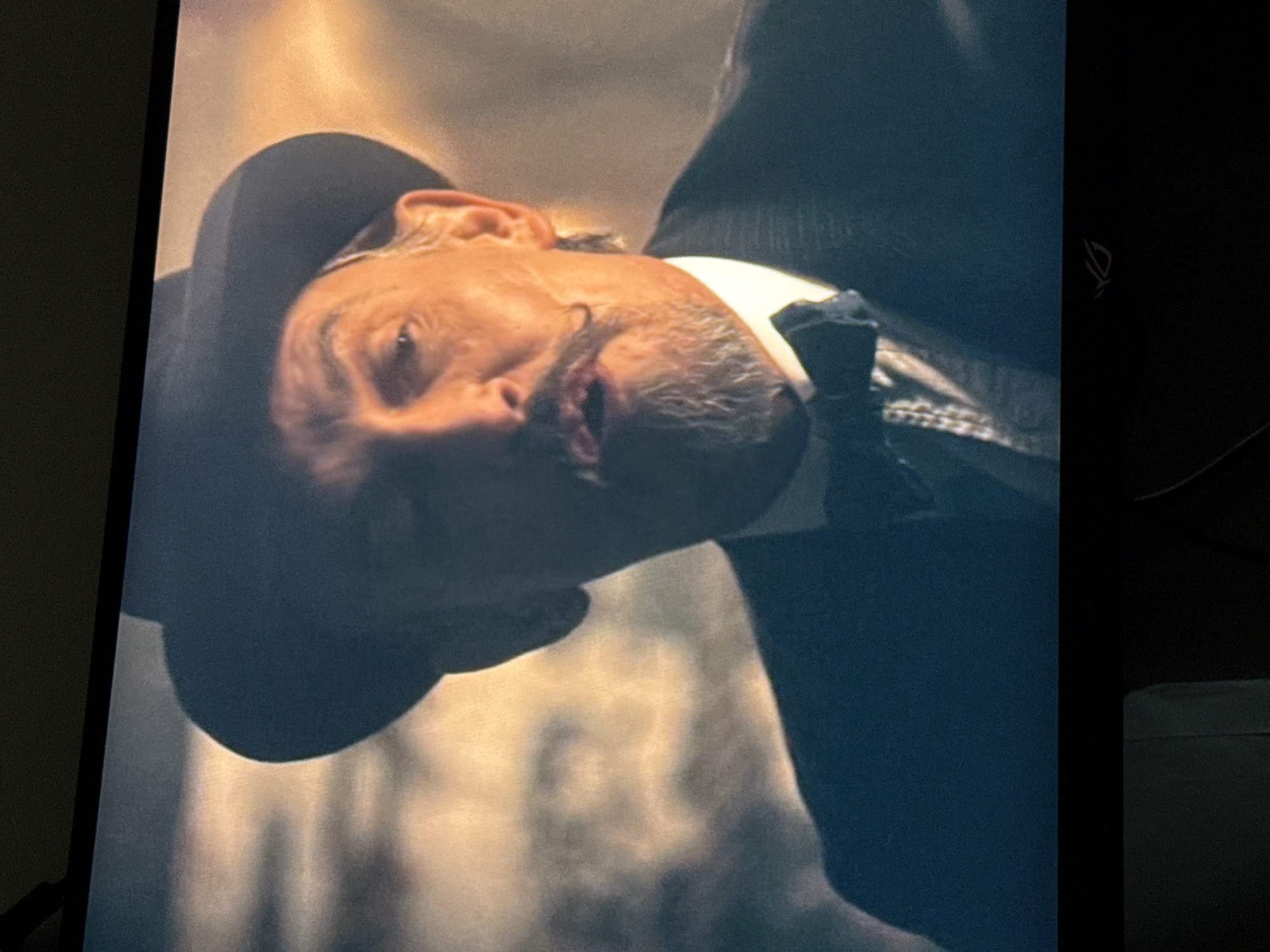 A low-angle, medium-close shot shows a man wearing a black hat and a dark suit. He has a mustache and a bow tie. The background is blurred and out of focus. The environment is unidentified, and the location in Unknown, Unknown, cannot be determined from the image.