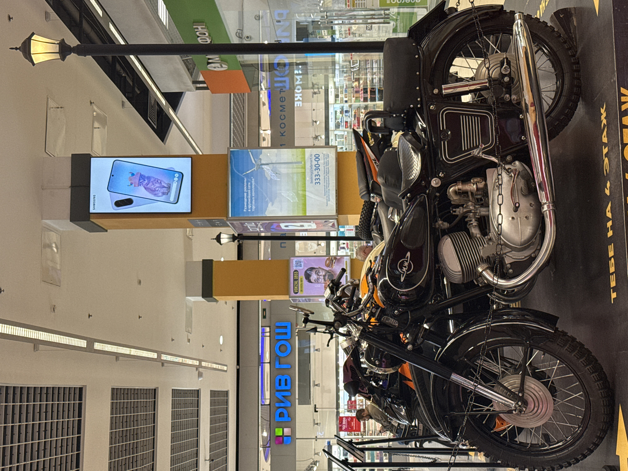 The image shows a vintage motorcycle display in what appears to be a shopping mall in Saint Petersburg, Russia. Several motorcycles are suspended by chains. The foreground features a black motorcycle with chrome accents. Behind the motorcycles, there are illuminated advertising signs and storefronts. The signs include ads for Samsung, phone number "333-30-00" for some sort of advertisement booking, the word "РИВ ГОШ" in bright blue, and "VOKZAL 1653." The background shows the interior of the shopping mall, including the ceiling, ventilation systems, and general retail environment. A sign in Russian reads "ТЕБЕ НА 4 ЭТАЖ" meaning "to you on the 4th floor." A street-style lamp post is visible.