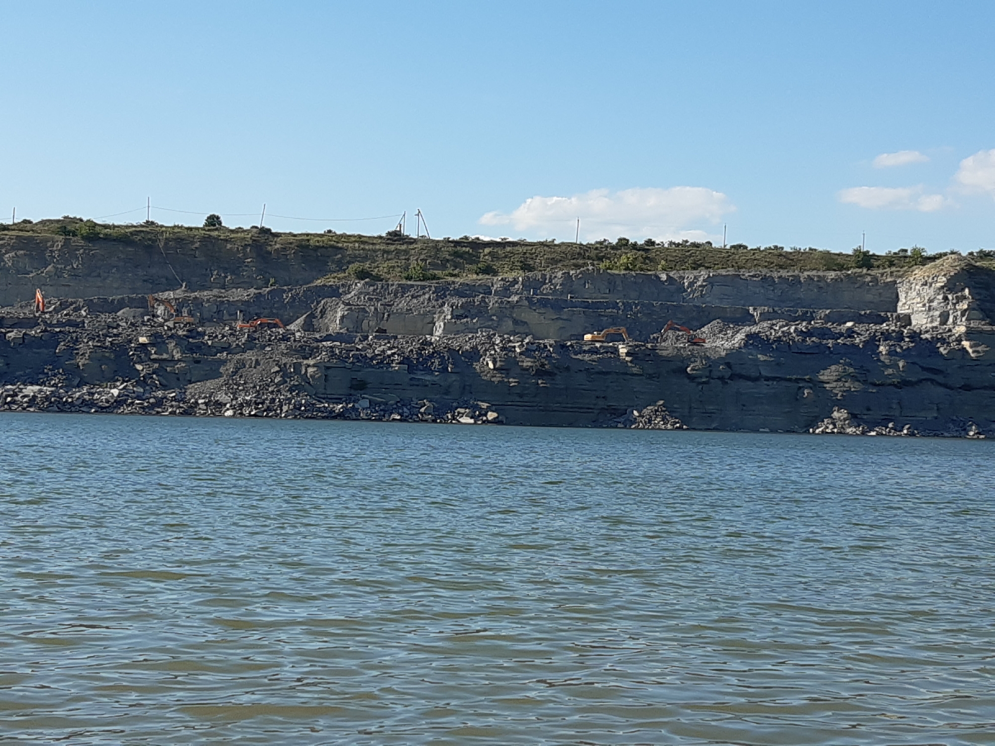 The image shows a quarry with excavators operating near a body of water. The quarry face is layered with rock, and piles of rubble are visible. Several orange excavators are actively working on the quarry floor, suggesting ongoing excavation or material handling. The sky is clear and blue with a few wispy clouds, indicating fair weather. The sun is high in the sky, suggesting it is daytime. The water in the foreground appears calm with gentle ripples, reflecting the sky. The overall scene conveys industrial activity within a natural landscape. No text is visible in the image.