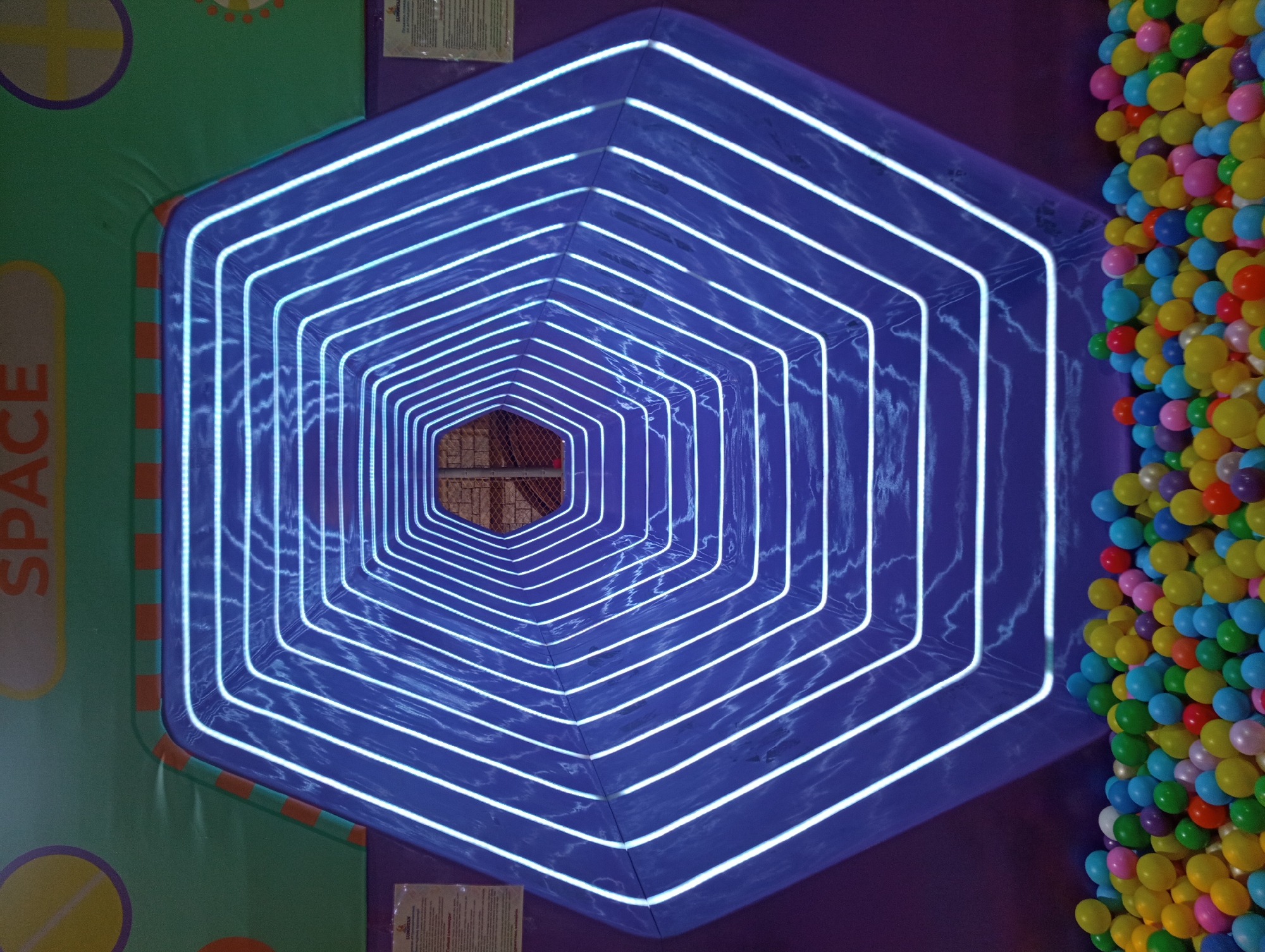 The image depicts a vibrant indoor play area, likely a children's amusement center, characterized by a tunnel with hexagonal framing illuminated by white LED lights. The tunnel entrance is a deep purple, and the outer wall displays the word "SPACE" in large, orange lettering against a green background adorned with circular graphics. The floor in front of the tunnel is filled with a large quantity of colorful plastic balls in various shades of red, yellow, green, blue, and purple. The overall scene suggests a playful and engaging environment. The location is identified as the "Limonad" amusement park in Almaty, Kazakhstan, as indicated by a visible sign in the upper left portion of the image.