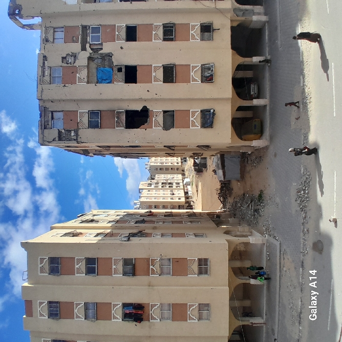 Rows of tan apartment buildings stand close together, some showing visible damage. One building has a blue tarp covering a hole. Windows appear dark or missing in several units. Debris litters the ground between the buildings. A street runs alongside the buildings, with several people walking along it. The sky is blue with scattered clouds. A 'Galaxy A14' watermark is visible in the bottom right corner.