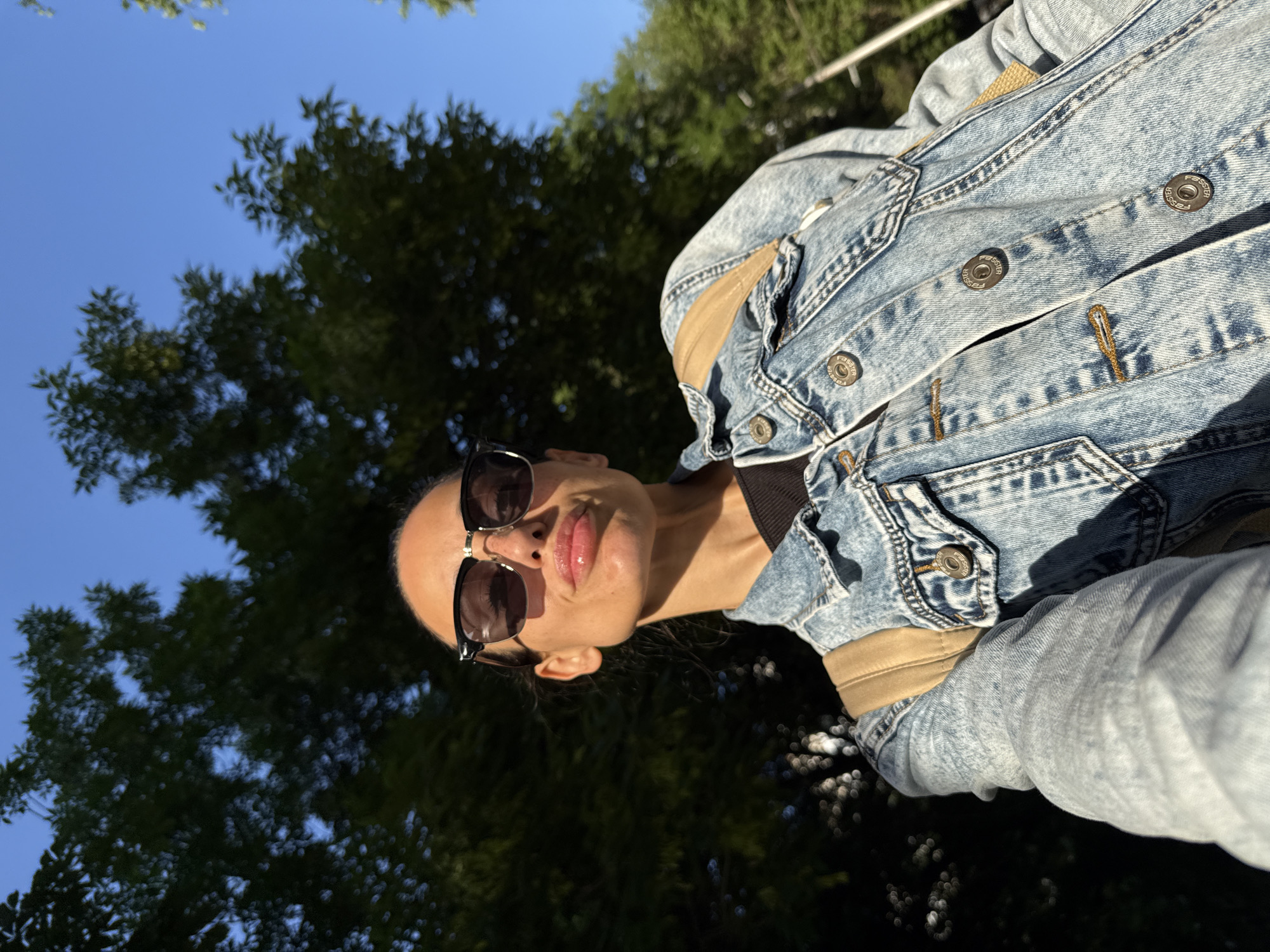 A woman wearing sunglasses and a denim jacket takes a selfie outdoors. The background includes a clear blue sky partially obscured by tree branches. The location is not identifiable as Pleven, Bulgaria based on the image content.