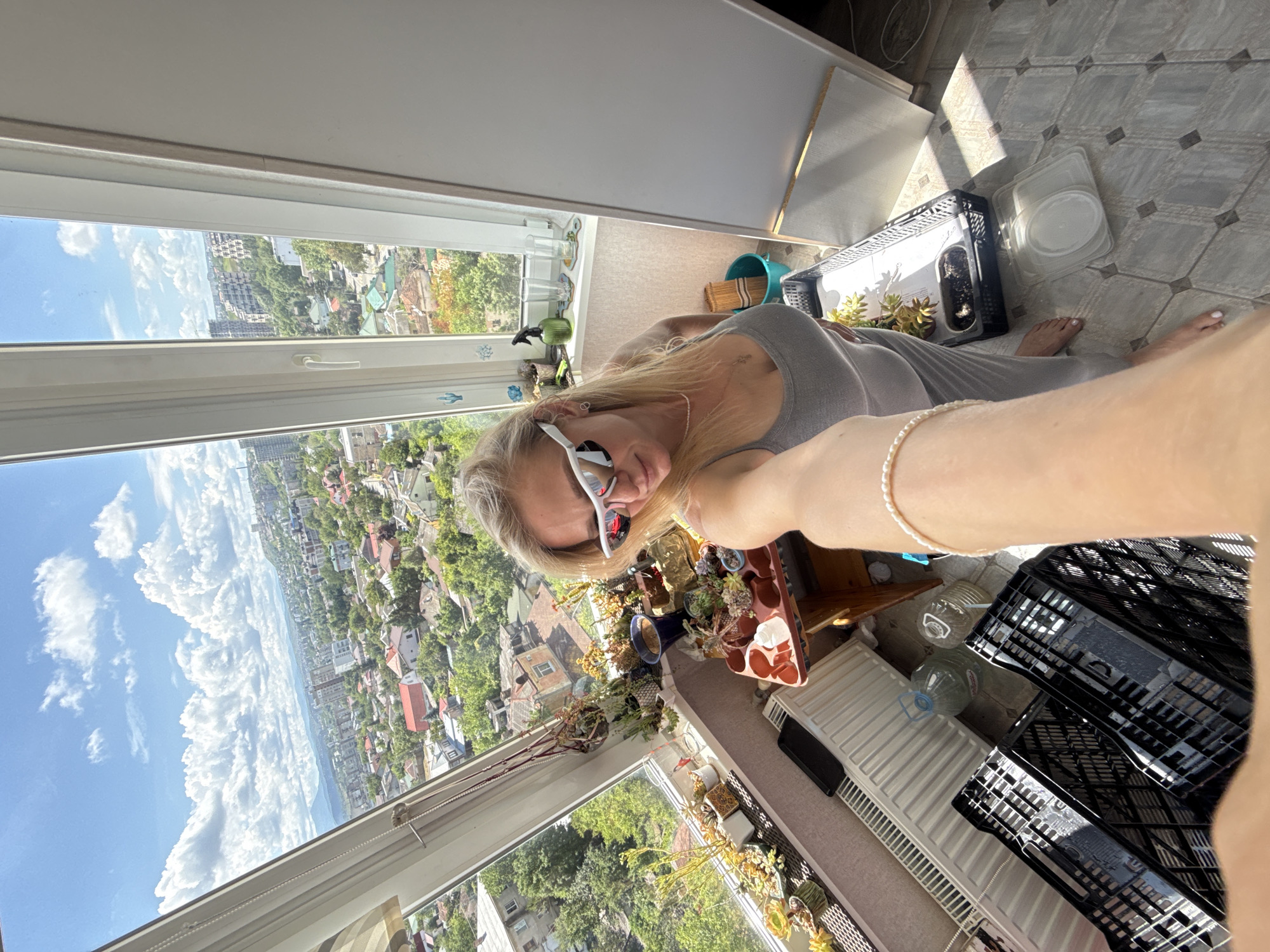 A woman with blonde hair, wearing white sunglasses and a grey tank top, is taking a selfie from a sunroom that overlooks a city. The room is filled with potted plants, and a radiator is visible below the window. In the foreground, a person's arm with a pearl bracelet is extended, holding the phone to take the photo. The city view outside the window shows numerous houses and buildings, with a sky full of clouds.