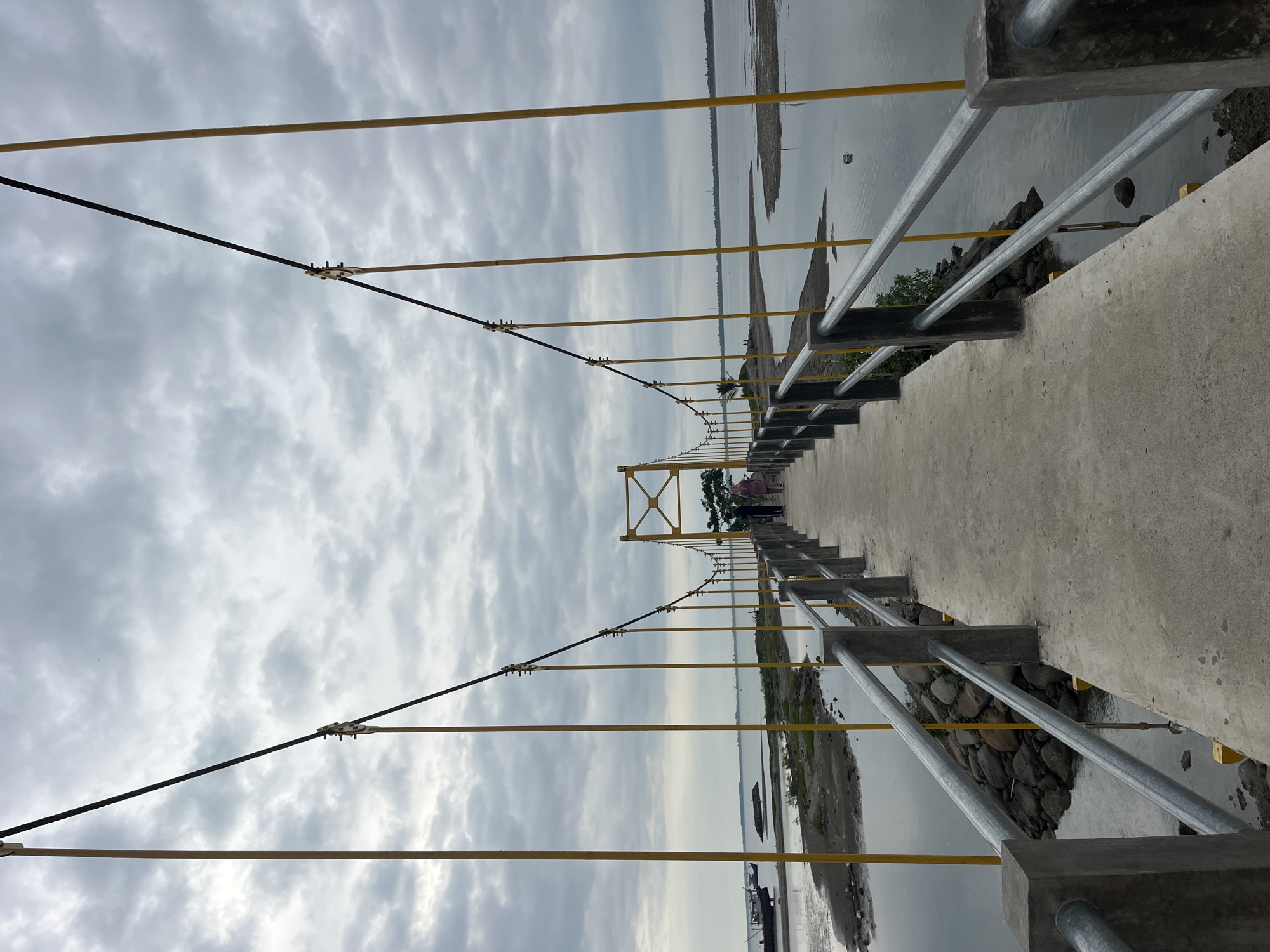 The image shows a suspension bridge spanning a body of water, likely a river or lake. The bridge has a concrete walkway and metal railings. Yellow vertical supports connect the walkway to the suspension cables above. The suspension cables are black and held in place by metal clamps. In the distance, the structure of the bridge is painted yellow. The sky is overcast with grey clouds. The water is murky. There are visible land formations on either side of the bridge. The location is likely within Lhokseumawe, Indonesia. In the distance, there are people walking on the bridge. Boats can be seen floating on the water.