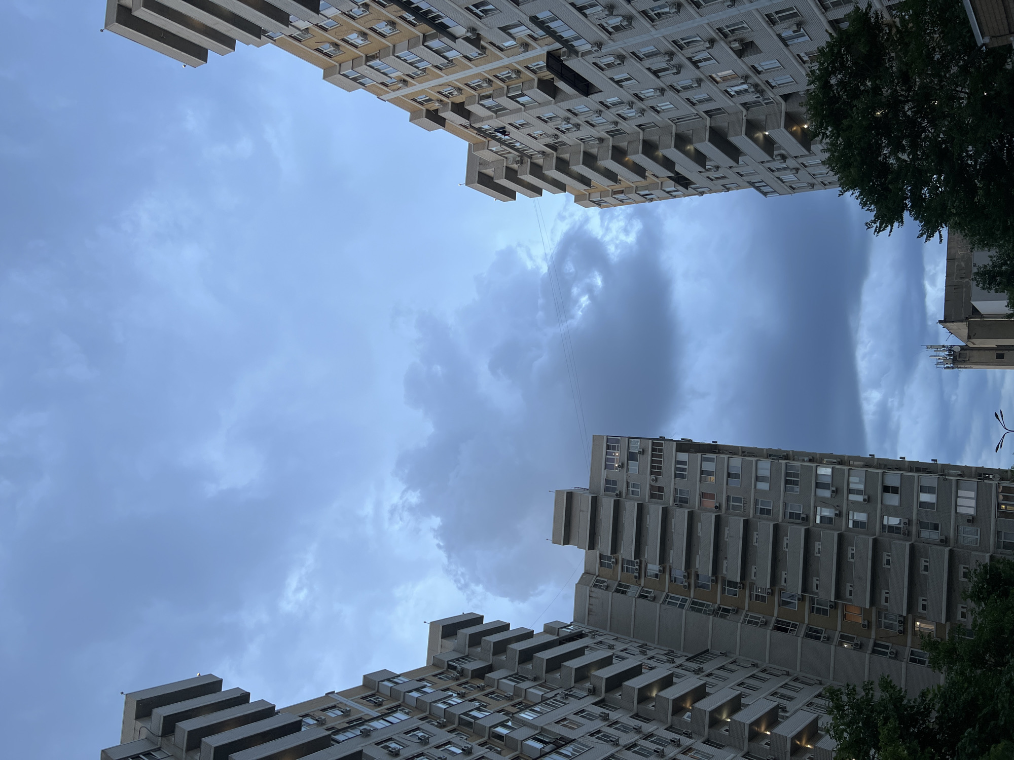 The image shows a view looking up at apartment buildings against a cloudy sky in Krasnodar, Russia. Two tall, multi-story apartment buildings dominate the scene, one on the left and one on the right, with their facades featuring numerous balconies and windows. The building on the left is lighter in color, appearing to be made of concrete and brick, while the building on the right is a similar construction but with some yellow brick accents. Below the buildings, the tops of dark green trees are visible. In the distance, a shorter building with an antenna is present. The sky is filled with dark, heavy clouds, suggesting overcast or potentially stormy weather. There are no people or animals visible in the frame. The overall impression is of an urban residential area under a dramatic sky.