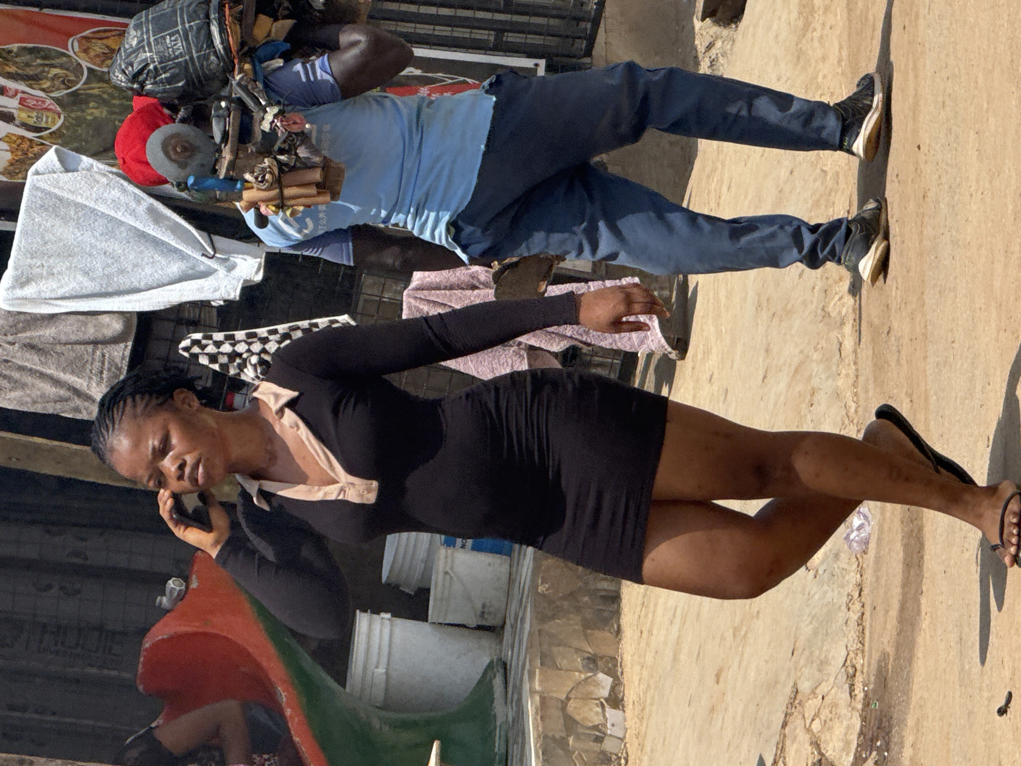 In an outdoor urban setting, likely in Abuja, Nigeria, a woman in a black, form-fitting dress with a collared neckline is walking and talking on a mobile phone. She is wearing black flip-flops. Behind her, a person in a blue t-shirt and dark blue pants is carrying a load of items on their back, secured with straps. The load includes what appears to be tools, including a grinder, and various bundles wrapped in fabric and plastic. The ground is dusty and unpaved. In the background, there are white buckets, a red and green structure, and some hanging laundry. A poster with images of food and drink is visible on a wall.