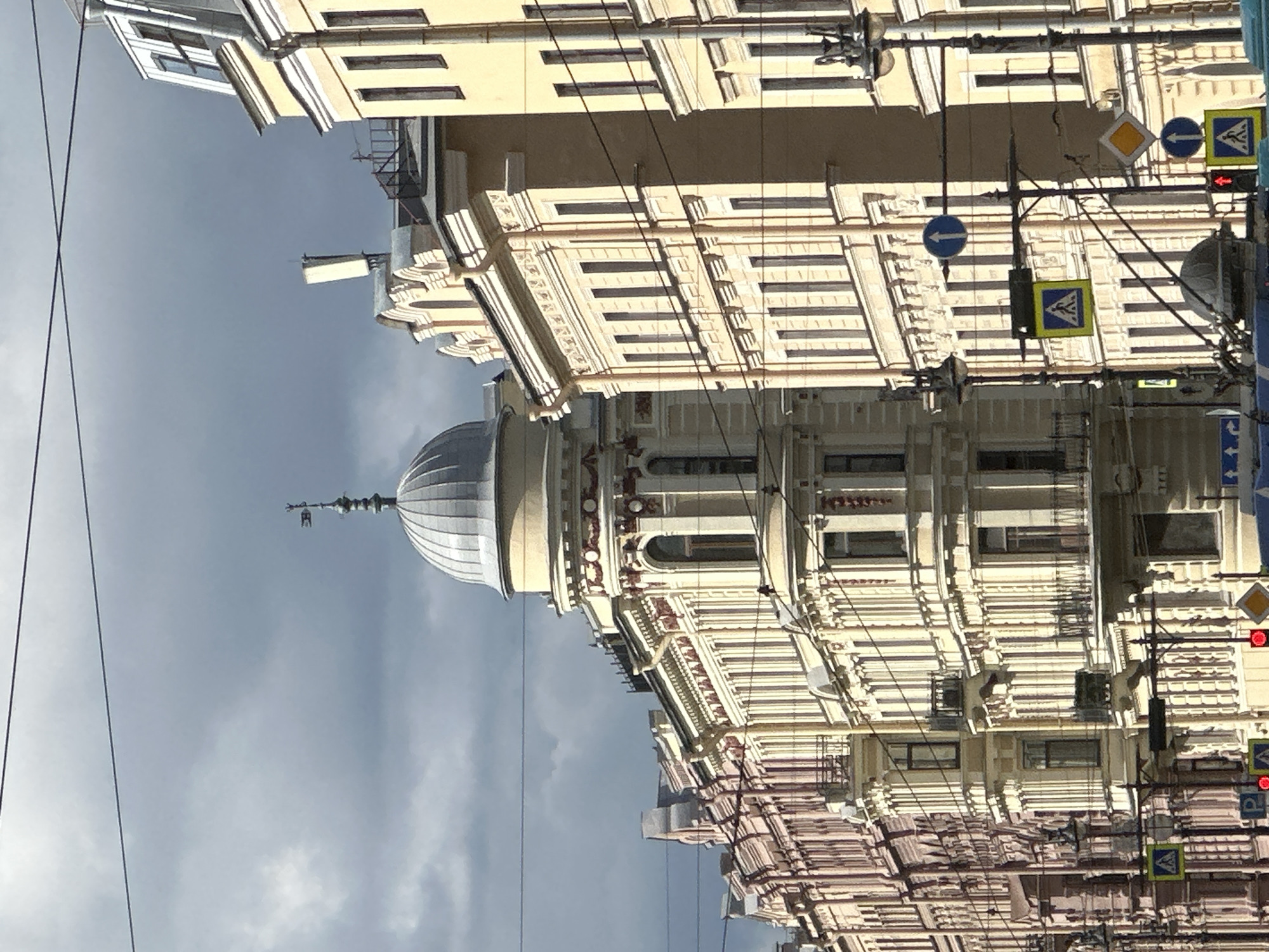 This image captures a street view in Saint Petersburg, Russia, focusing on the architectural grandeur of its historic buildings under a dramatic, overcast sky. The buildings are predominantly tall, multi-story structures with ornate facades, characterized by classical architectural elements such as columns, decorative moldings, and arched windows. The style suggests a pre-revolutionary era, likely late 19th or early 20th century.
On the left, a row of buildings exhibits a more muted, possibly faded color palette, with subtle detailing. As the eye moves to the right, the buildings become more prominent and brightly lit, featuring a creamy yellow hue. One building stands out with a distinctive dome, topped by a weather vane, adding a classical civic or religious touch to the urban landscape. The windows are numerous and arranged in a regular pattern, with some featuring ornate balconies.
The scene is also populated with urban infrastructure. Overhead, a network of power lines or communication cables crisscrosses the sky, a common feature in many cities. Below, traffic signals and pedestrian crossing signs are visible, indicating a busy thoroughfare. Specifically, there are yellow square signs with pedestrian symbols and blue circular signs with upward-pointing arrows. Red traffic lights are also present. The presence of these signs and the perspective from street level suggest the viewpoint is from a sidewalk or the street itself.
The sky is a significant element, with dark, heavy clouds dominating the upper portion of the frame, contrasting with patches of sunlight that illuminate the buildings. This creates a dynamic interplay of light and shadow across the facades. The overall impression is one of a dignified and historically rich urban environment, characteristic of Saint Petersburg's famed architectural heritage, even under less-than-ideal weather conditions. No individuals or overt actions are clearly discernible in the frame, emphasizing the architectural and environmental aspects of the scene.