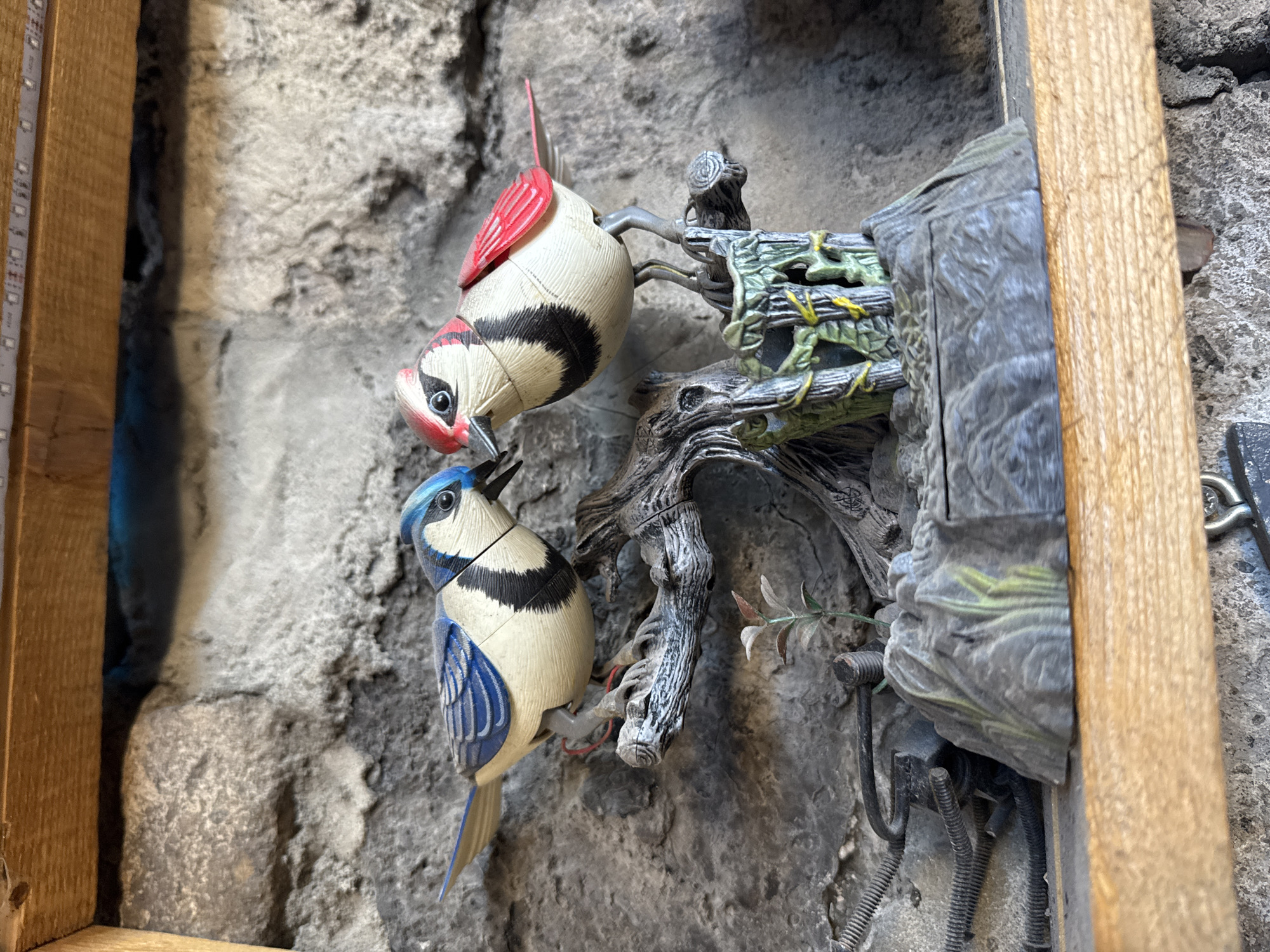 This close-up image displays a decorative scene featuring two artificial bird figures attached to a gnarled, dark-colored branch-like structure. The background is a rough, textured grey wall, possibly stone or concrete. The entire setup appears to be recessed or framed by vertical wooden planks on the left and right sides.
The top bird figure has a predominantly white body with distinct black stripes on its back and wings, and vibrant red coloration on its head, chest, and tail. It is angled downwards, as if pecking or looking towards something below. Below it, the second bird figure also has a white body with black stripes, but features bright blue on its head, wings, and tail, resembling a blue jay. It is also angled downwards. Both birds are firmly attached to the artificial branch structure.
The branch structure itself is intricate, appearing weathered with details resembling bark and possibly some green moss or vines. Adjacent to the branch, on the right side, is a dark grey, block-like structure with a textured surface, possibly mimicking a rock or a small, enclosed dwelling. Several wires and mechanical components are visible beneath and around this structure, suggesting the birds might be part of an animated display, such as a cuckoo clock or a similar automata.
On the inner edge of the left wooden frame, a strip of small lights, likely an LED strip, is visible, casting artificial light into the display. Faint, illegible markings are visible on this strip. The lighting is bright and even, indicating an indoor setting. The image does not provide specific cues regarding the time of day, weather, or emotions, but the overall impression is of a meticulously crafted, static display. The location context provided is Yerevan, Armenia.