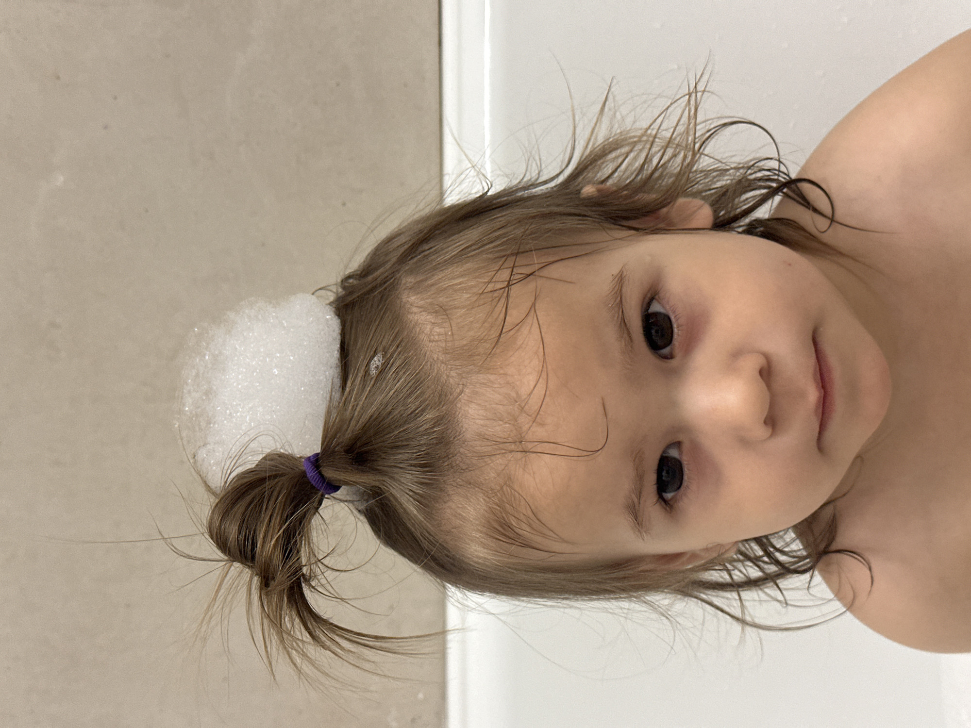 A young child with light brown hair and dark eyes looks into the camera with a slightly tilted head. Her hair is partially pulled back into a small ponytail with a purple hair tie on the upper left side of the frame, topped with a mound of white soap bubbles. The child's skin is fair. The background suggests a bathroom setting with a beige wall and a white bathtub edge. The scene appears to be a candid shot of a child during bath time.