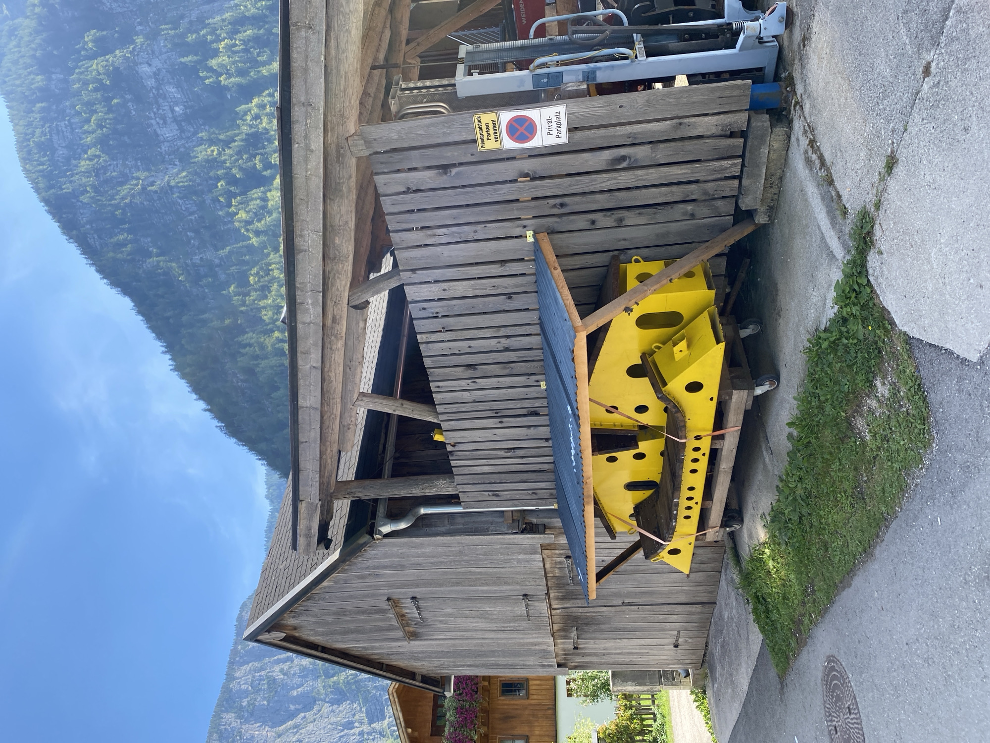 The image captures a scene in Hallstatt, Austria, featuring a weathered wooden barn and an attached shelter. Underneath the shelter, a large, bright yellow, industrial-looking object is mounted on a wheeled platform. This object appears to be a piece of machinery or equipment, possibly related to a mine or industrial operation, given Hallstatt's historical mining background. The object is secured with straps and is partially covered by a dark roof. A wooden fence stands to the right of the shelter, with a sign indicating "Privatgrundstück Parken Verboten!" which translates to "Private property, parking forbidden!". Behind the fence, a part of a light blue and gray forklift is visible. The barn itself is constructed of vertical wooden planks and has a sloped, dark-colored roof. In the background, the imposing, tree-covered mountains characteristic of the Hallstatt region rise under a clear blue sky. The ground in front of the barn is a mix of concrete and asphalt.